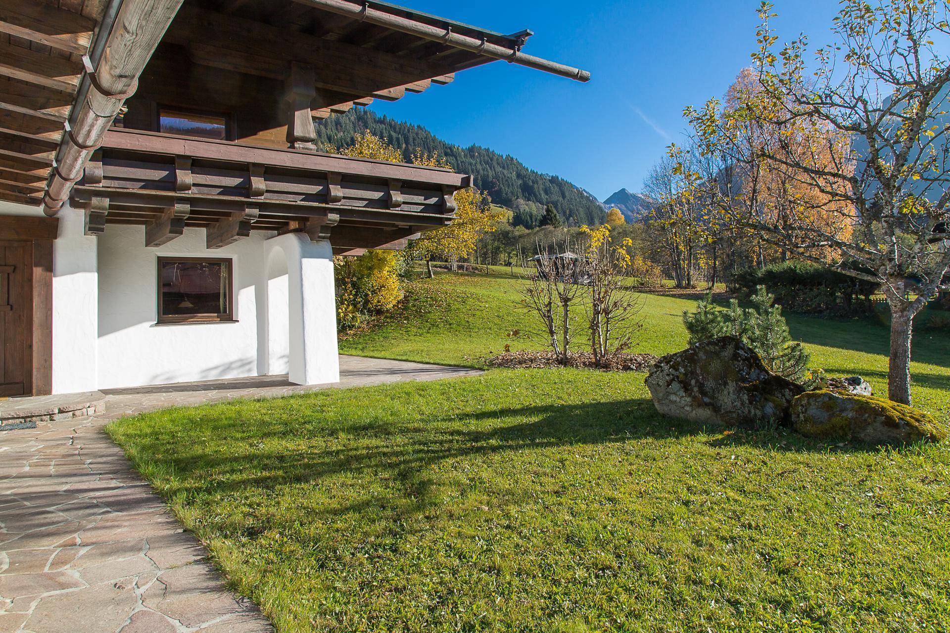 Sonnige Gartenansicht eines Hauses im alpinen Stil mit grüner Wiese und herbstlichen Bäumen.