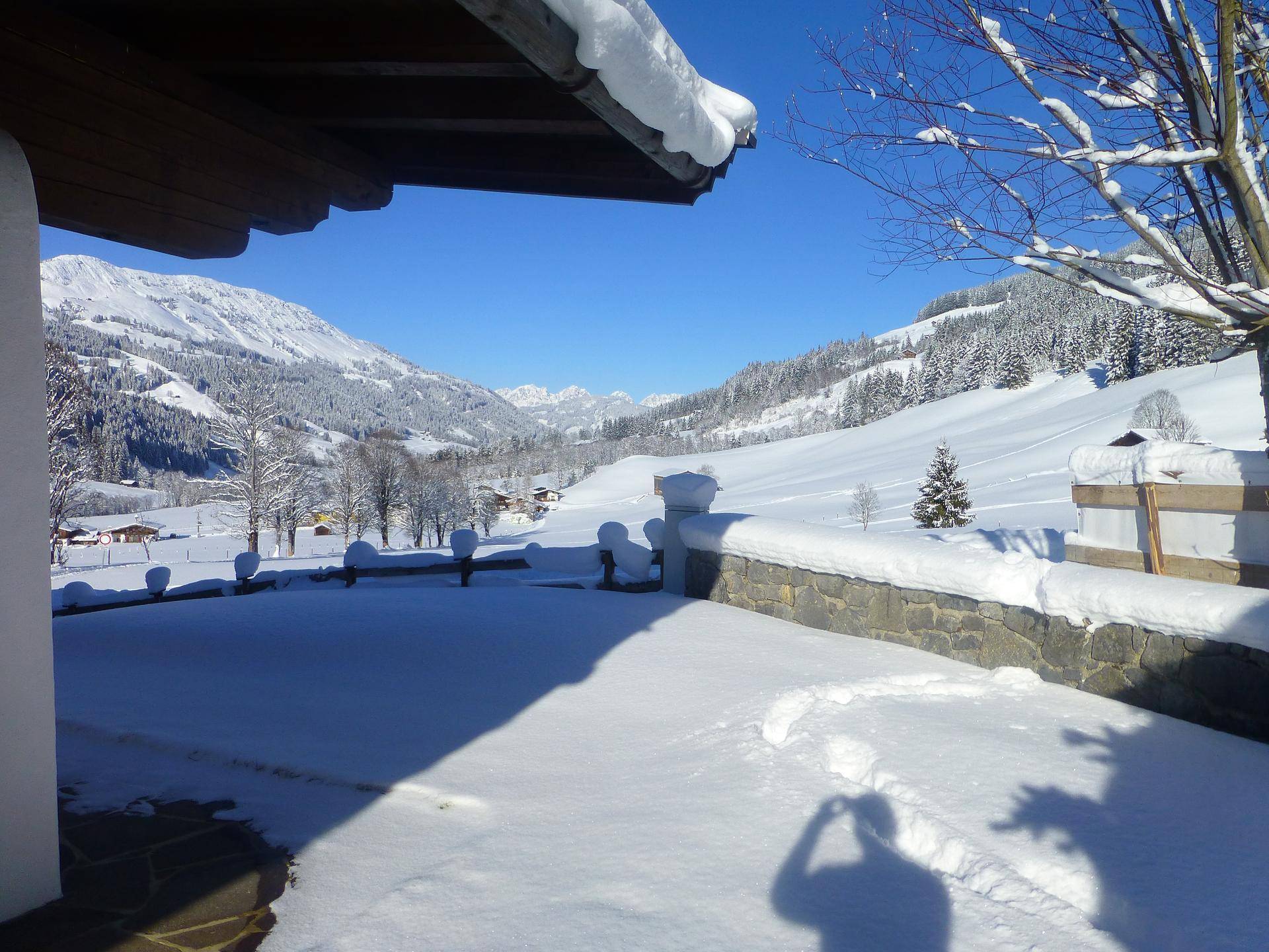 Verschneite Terrasse mit Steinmauer und weitem Blick auf die winterliche Berglandschaft unter blauem Himmel.