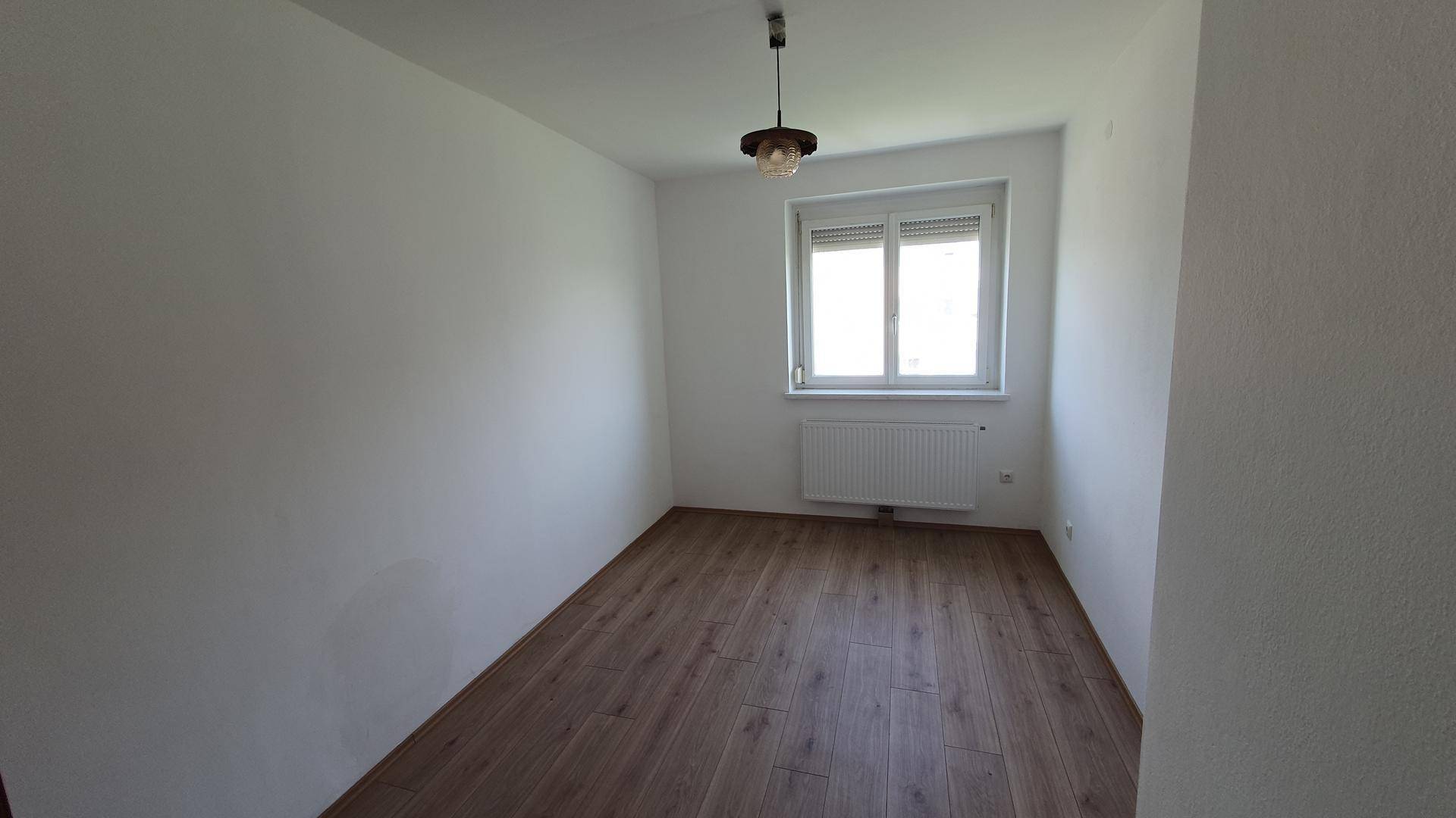 Geräumiges Schlafzimmer mit Holzboden, weißen Wänden und einem Fenster mit Rollladen, bereit zur Einrichtung.