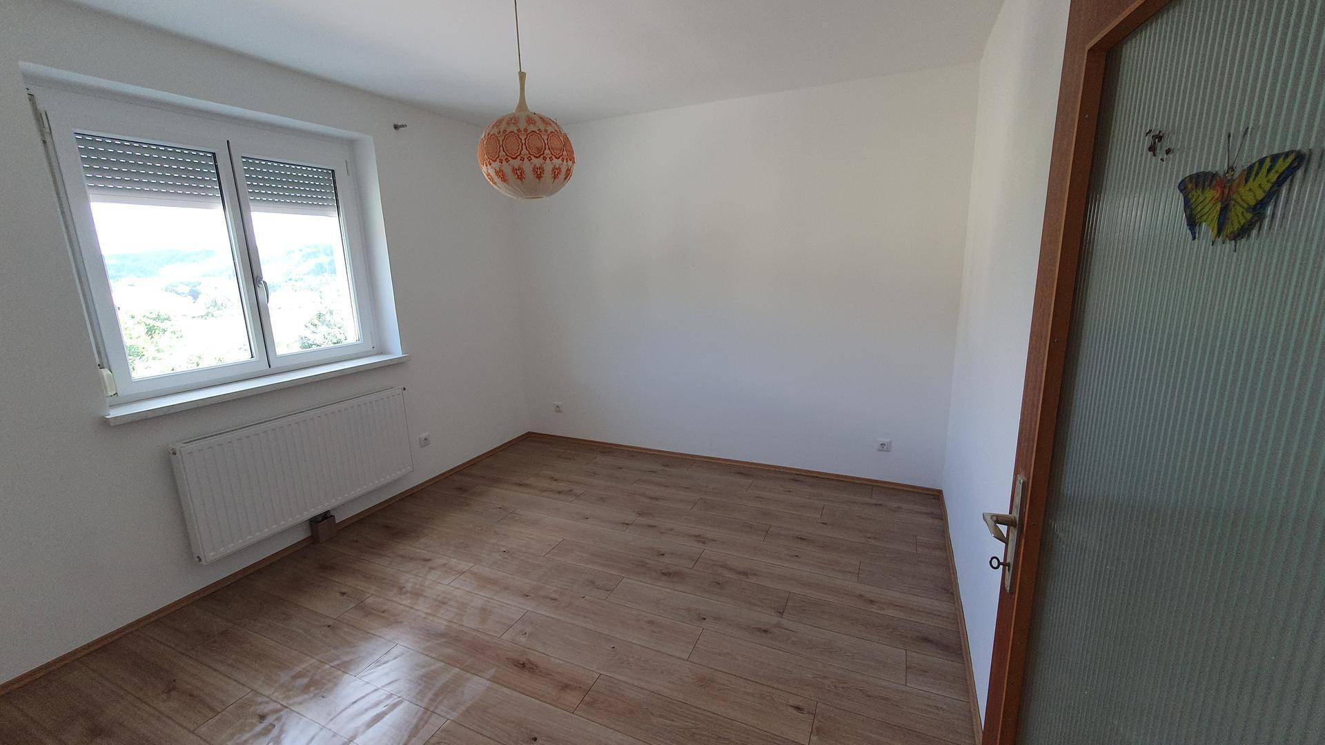 Leeres Schlafzimmer mit Holzboden und Fenster, das einen Blick ins Grüne und viel Tageslicht bietet.
