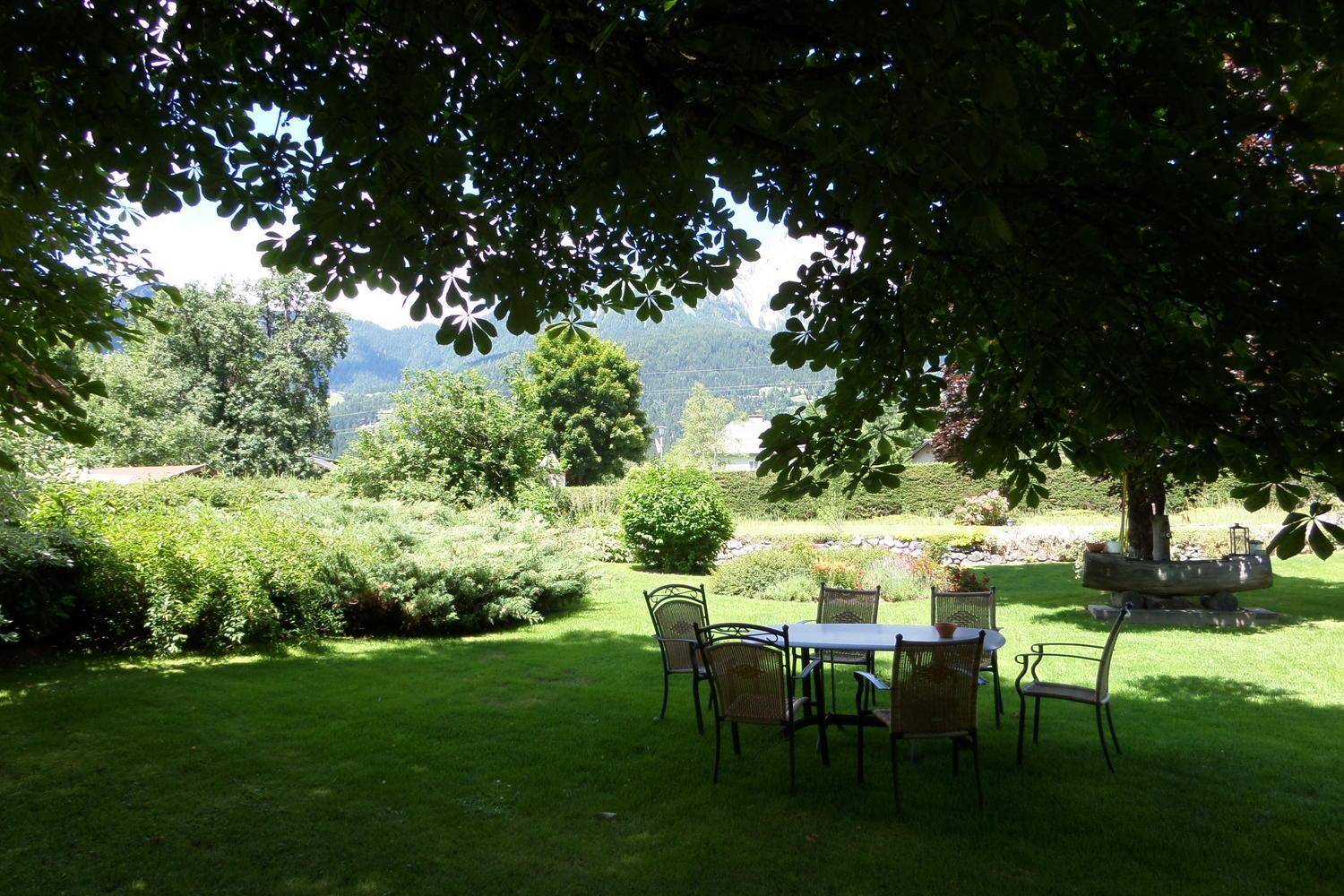 Schattiger Gartenbereich mit Sitzgruppe unter einem Baum, umgeben von Rasen und Bergblick.