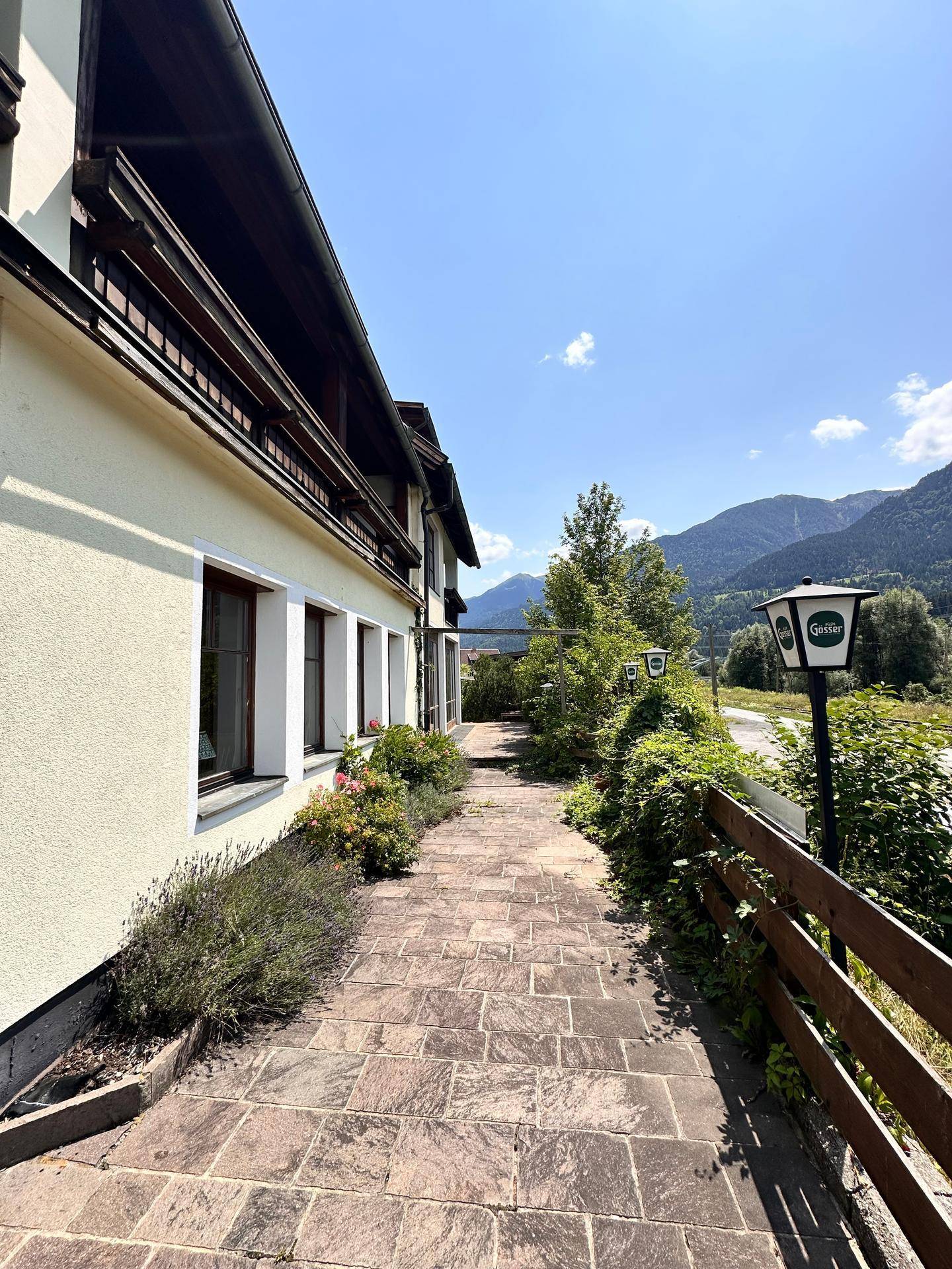 Langer, gepflasterter Weg entlang der Gebäudeseite mit Blick auf die umliegende Berglandschaft.