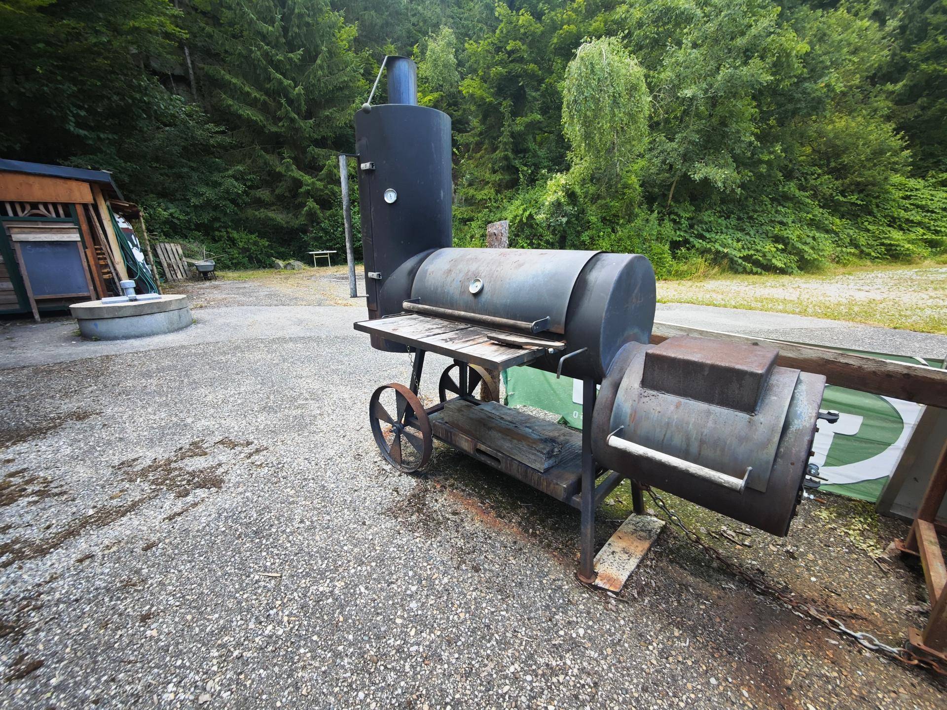 Großer, robuster Barbecue-Smoker auf einem Kiesplatz, umgeben von üppiger Vegetation.
