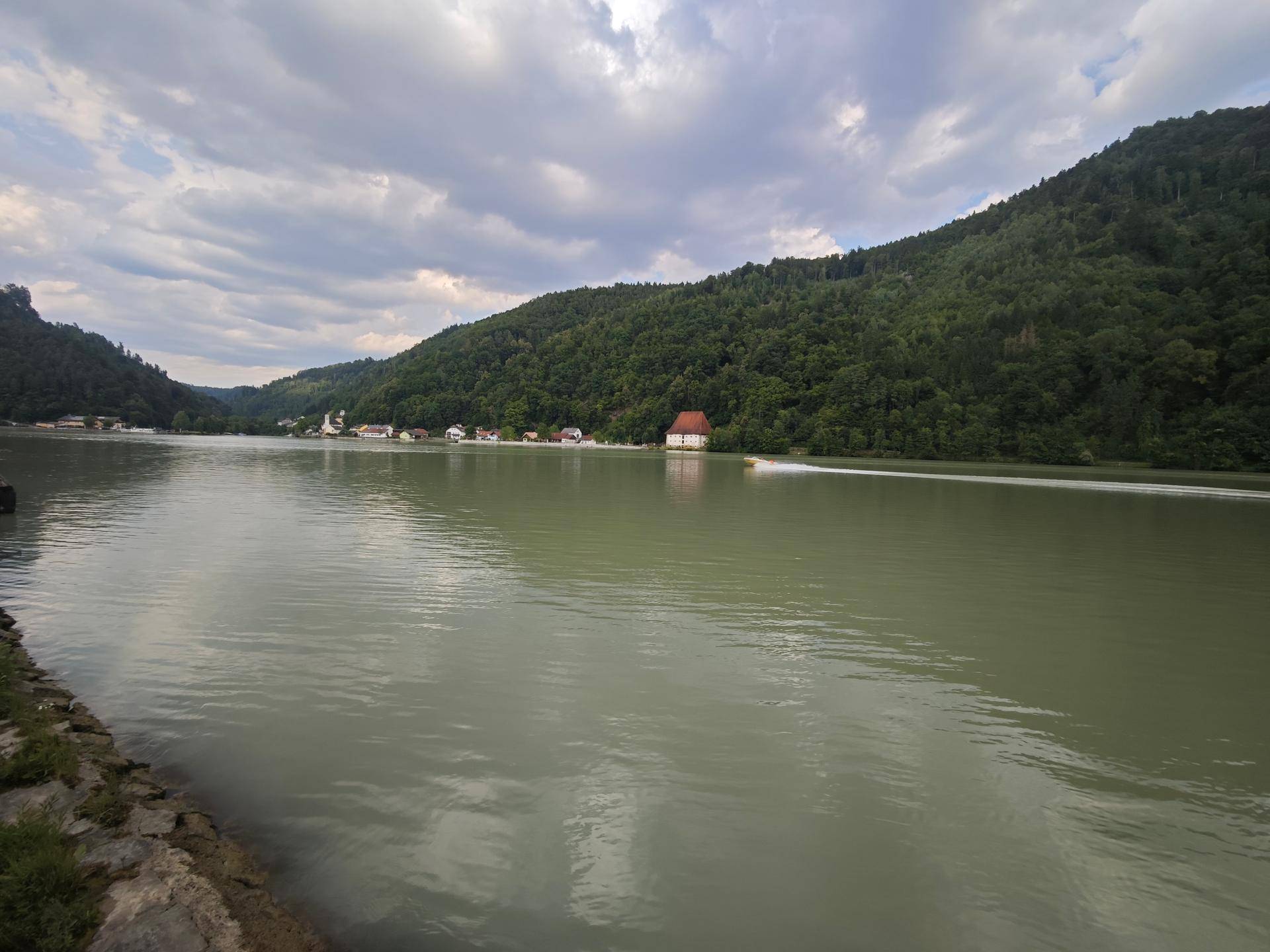 Weitläufiger Blick auf einen ruhigen Fluss oder See, umgeben von bewaldeten Hügeln und vereinzelten Gebäuden.
