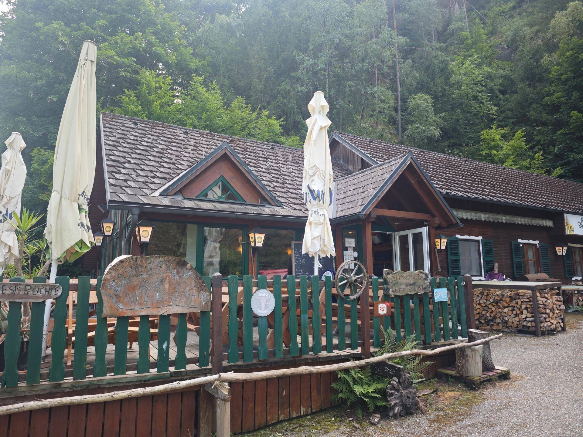 Charmantes Gasthaus mit großer Außenterrasse, Sonnenschirmen und rustikalem Holzzaun, umgeben von Wald.