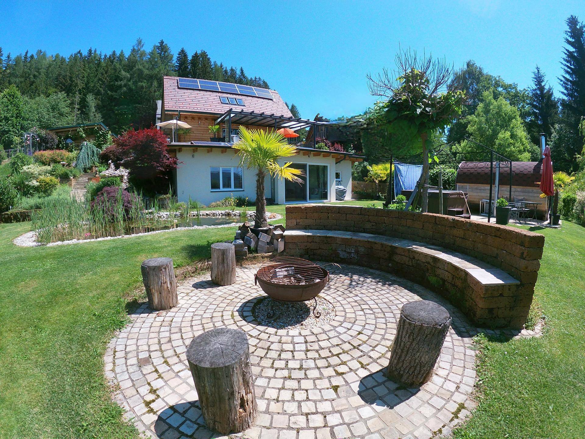 Gemütliche Feuerstelle mit runder Sitzbank und Holzstämmen im weitläufigen Garten mit Bergblick.
