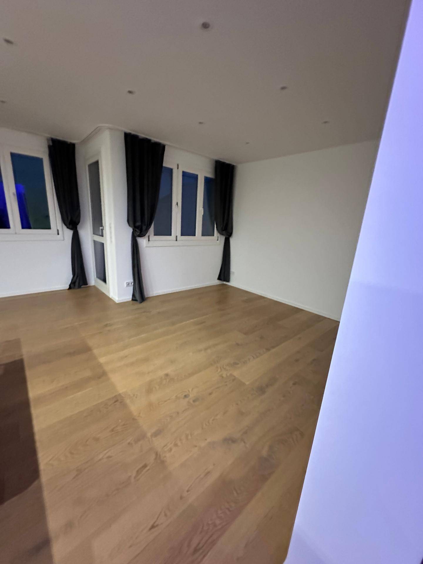 Heller Wohnraum mit Holzboden, mehreren Fenstern und dunklen Vorhängen.