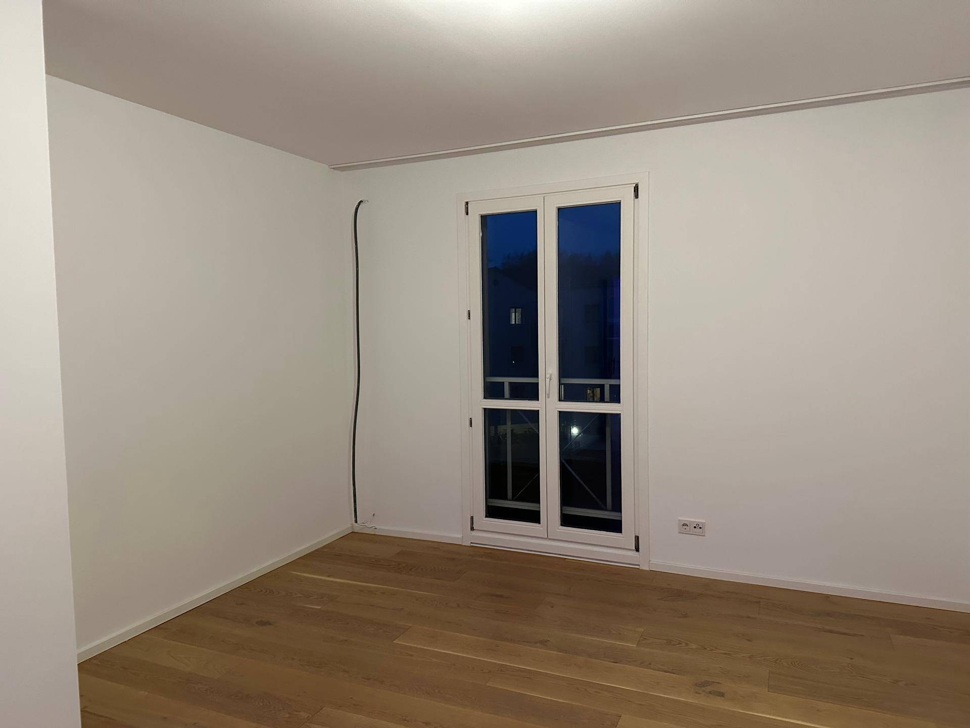 Leeres Zimmer mit Holzboden und französischem Balkonfenster am Abend.