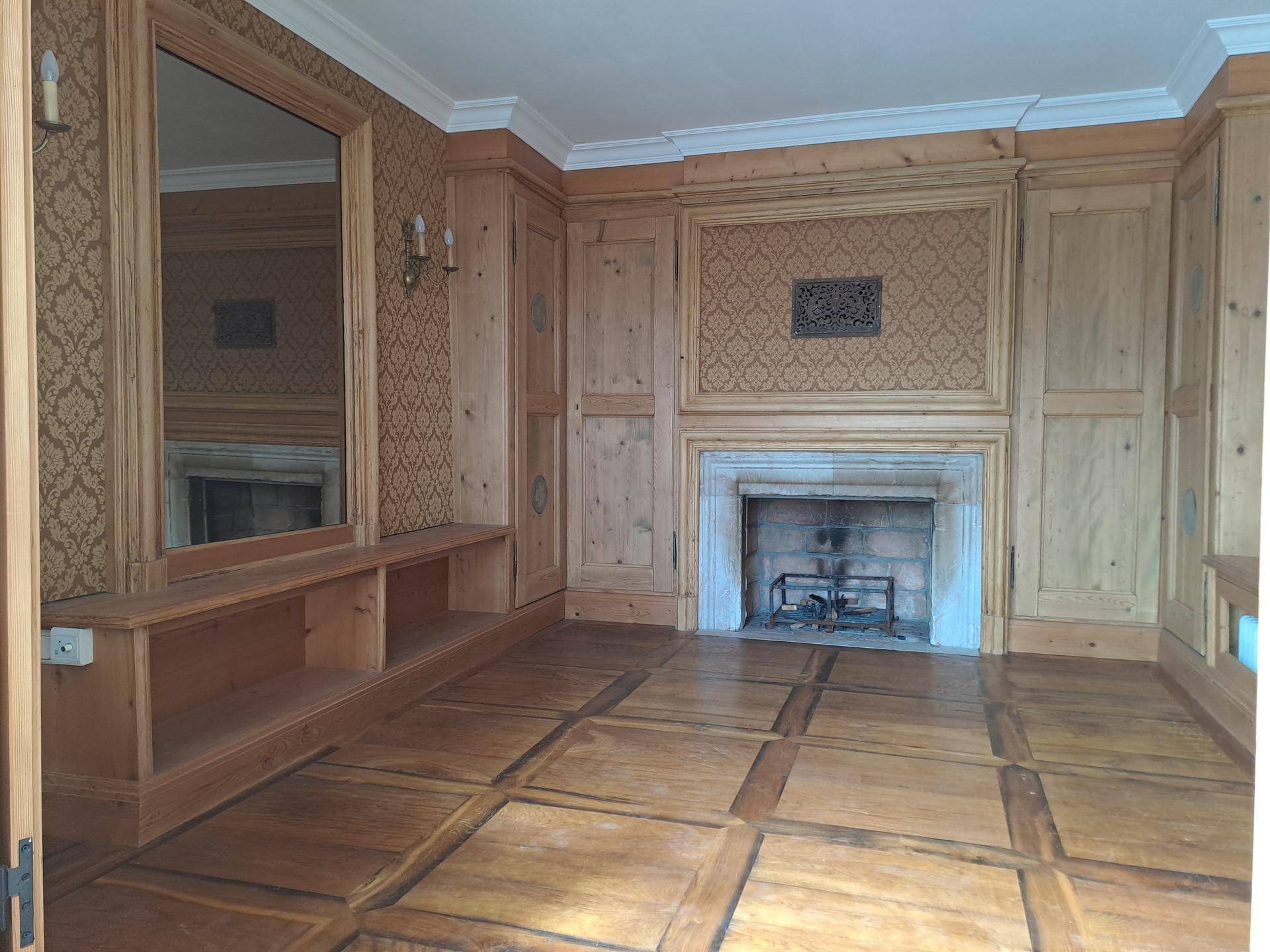 Traditionelles Zimmer mit Holztäfelung, Kamin und gemusterten Tapeten in warmen Farbtönen.