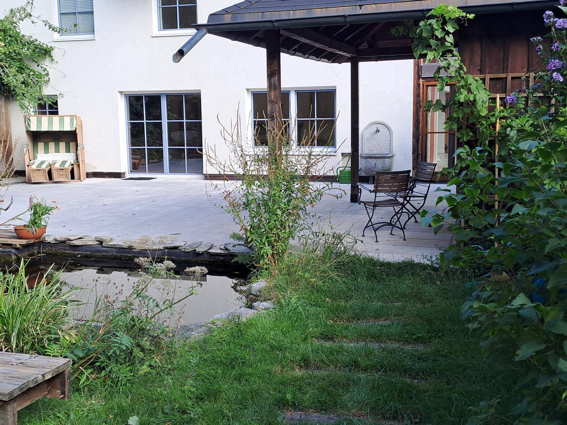 Großer Garten mit Teich, gepflasterter Terrasse und Blick auf die weiße Hausfassade.