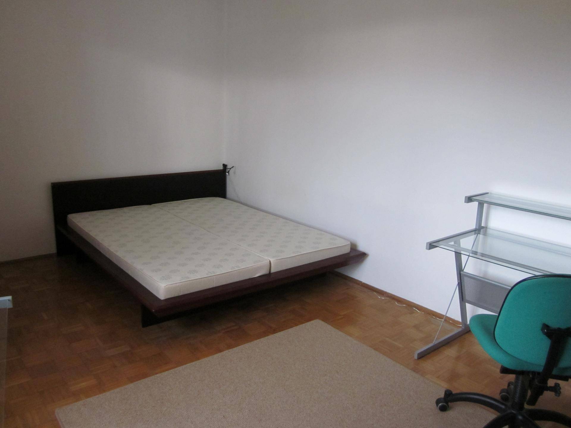 Einfaches Schlafzimmer mit Doppelbett, Schreibtisch und Parkettboden, bietet eine funktionale und helle Wohnatmosphäre.