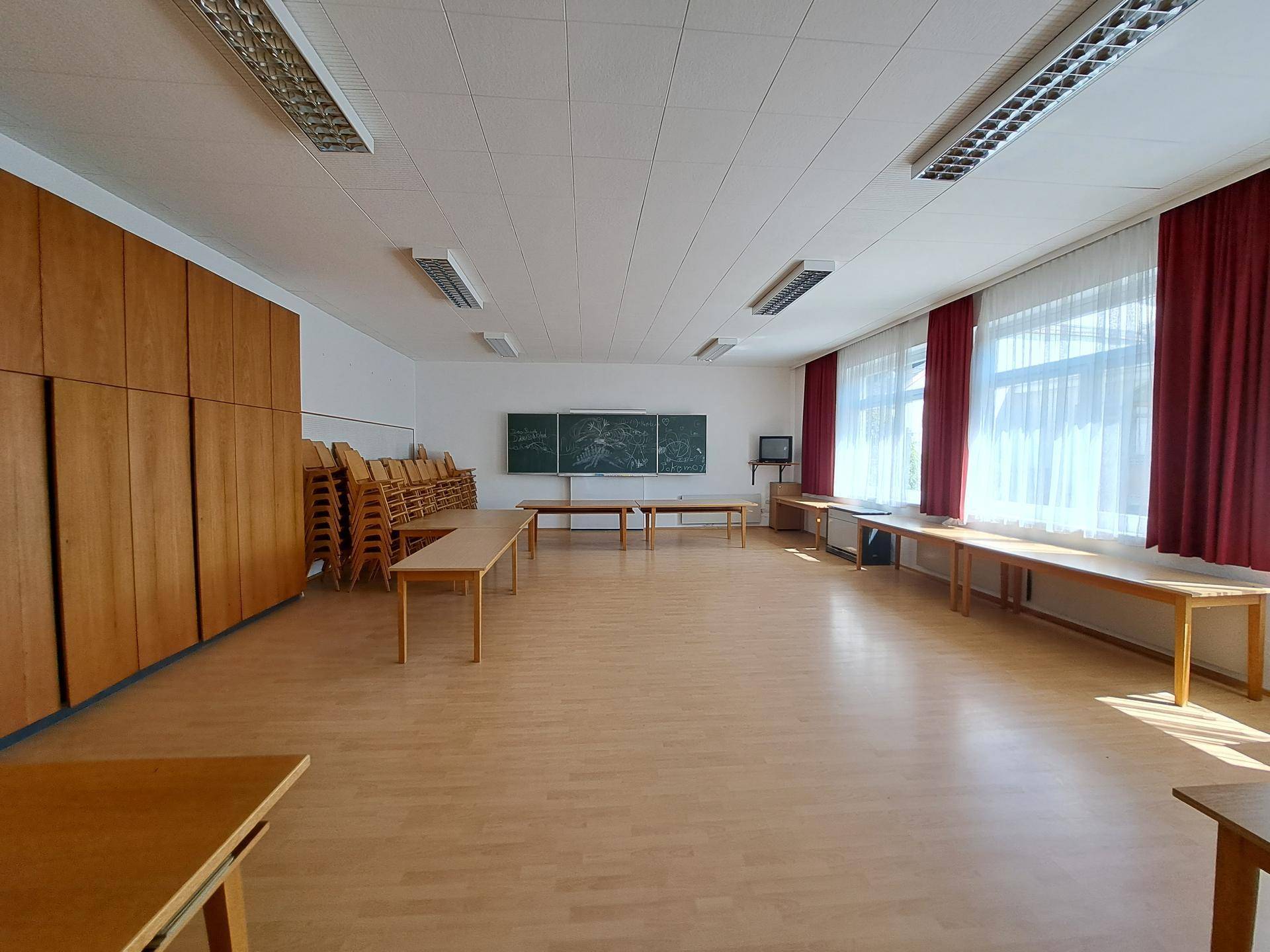 Heller Mehrzweckraum mit Holztischen, Schränken, Tafel und großen Fenstern mit roten Vorhängen.