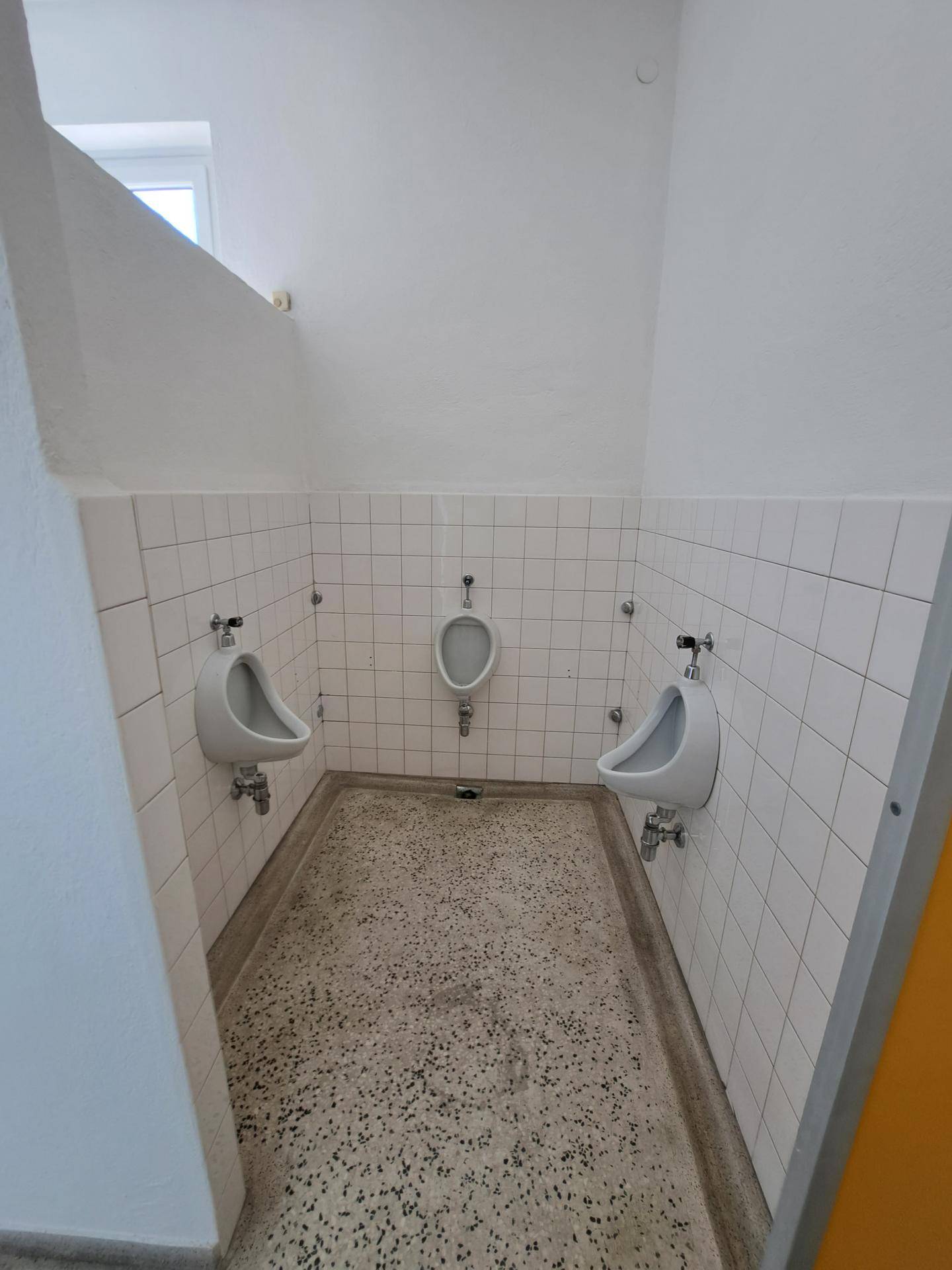 Öffentliche Toilettenanlage mit mehreren Urinalen, weißen Fliesen und Terrazzoboden.