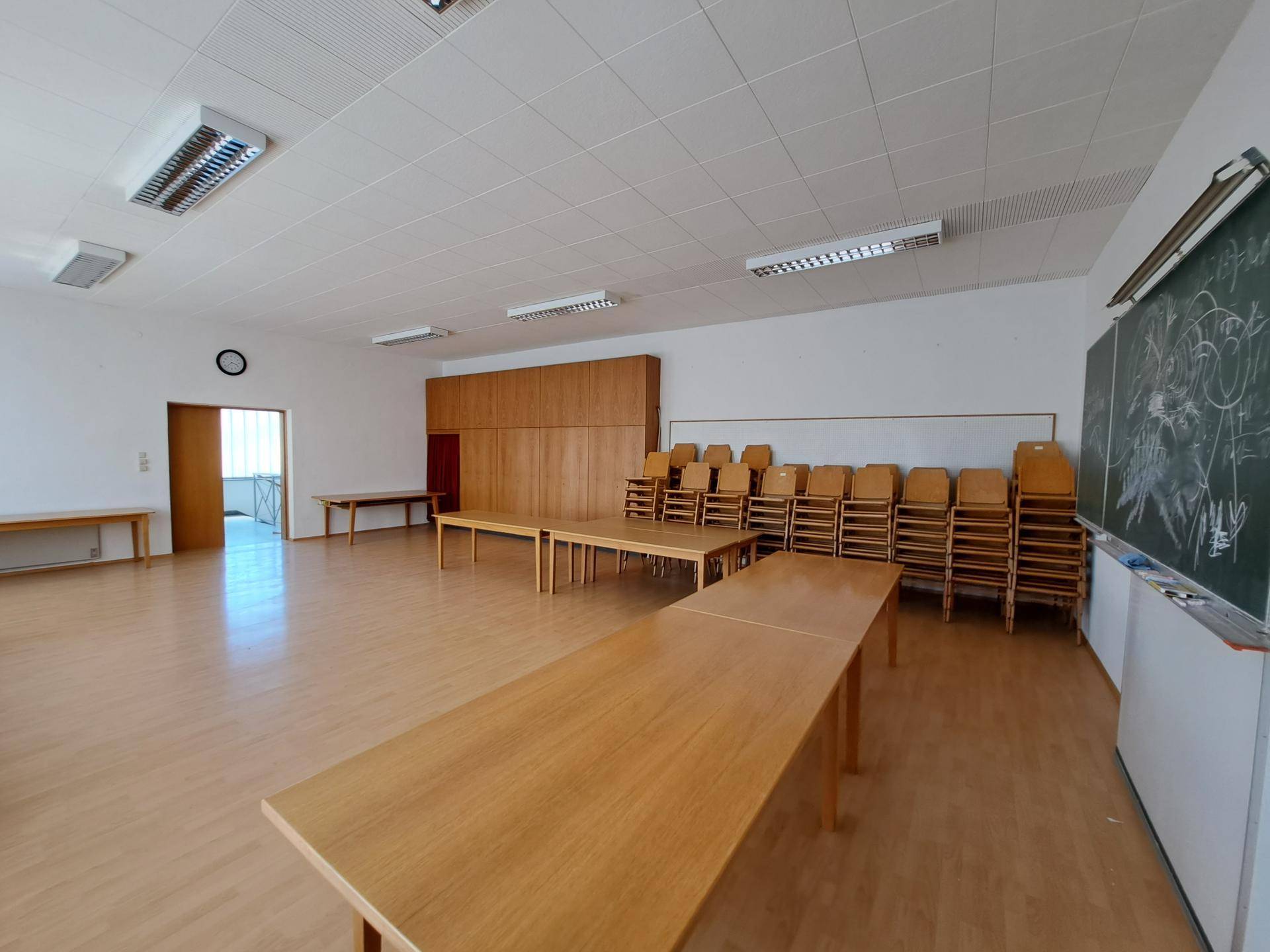Geräumiger Mehrzweckraum mit gestapelten Stühlen, einer Tafel und Holzschränken an der Wand.