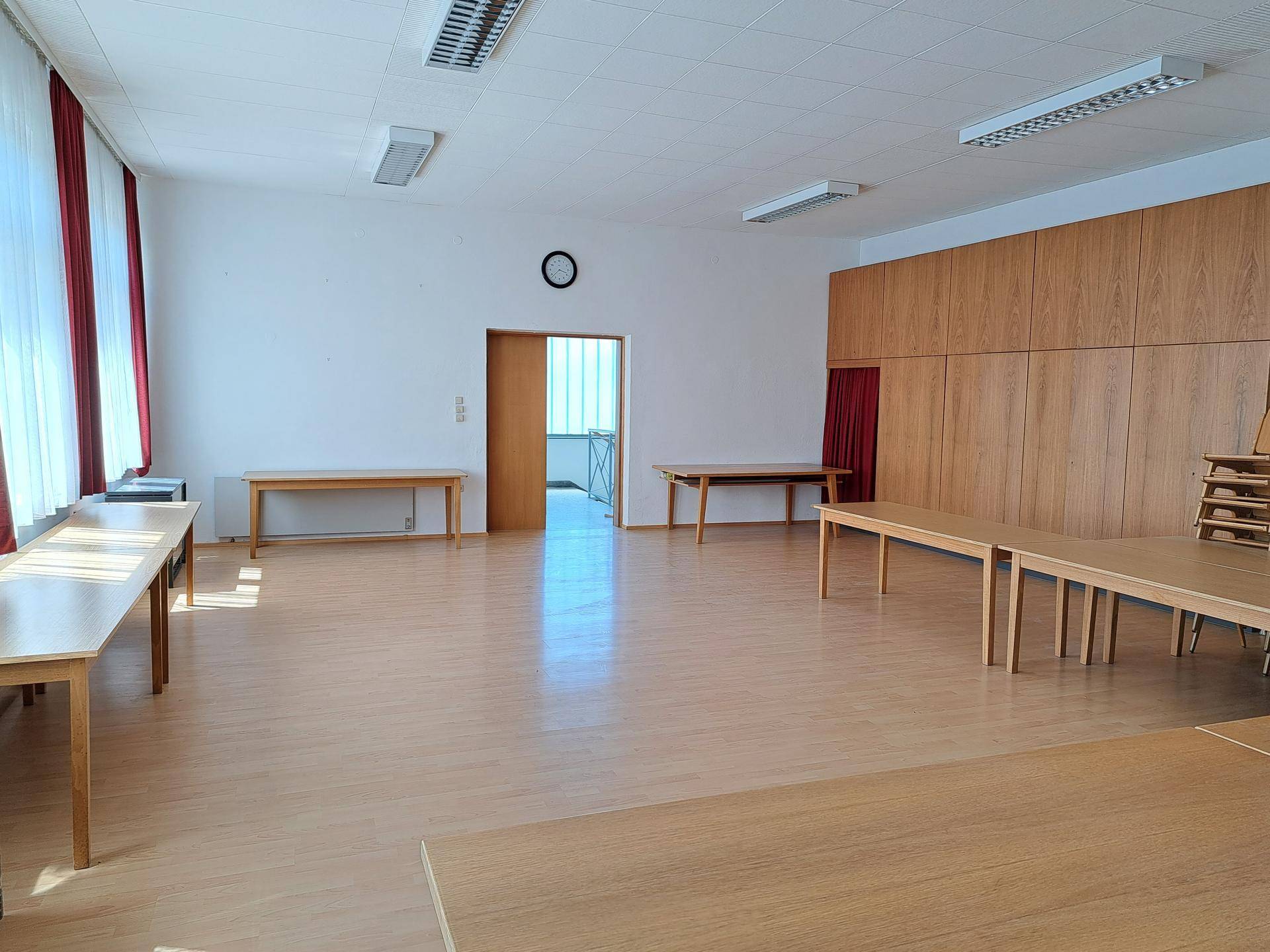 Großer, heller Mehrzweckraum mit Holztischen, Parkettboden und roten Vorhängen an den Fenstern.