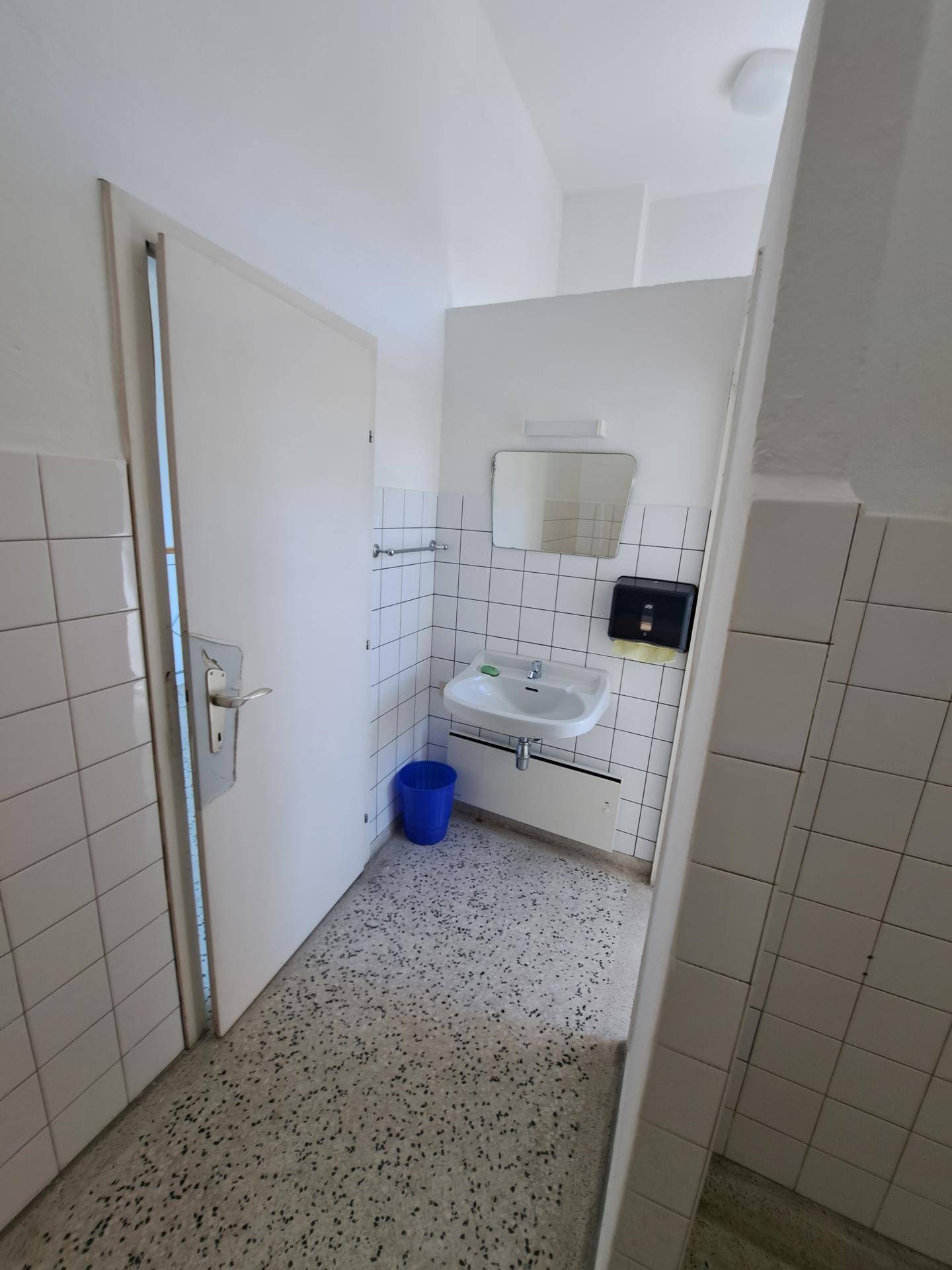 Einfacher WC-Raum mit Waschbecken, weißen Fliesen und Terrazzoboden.