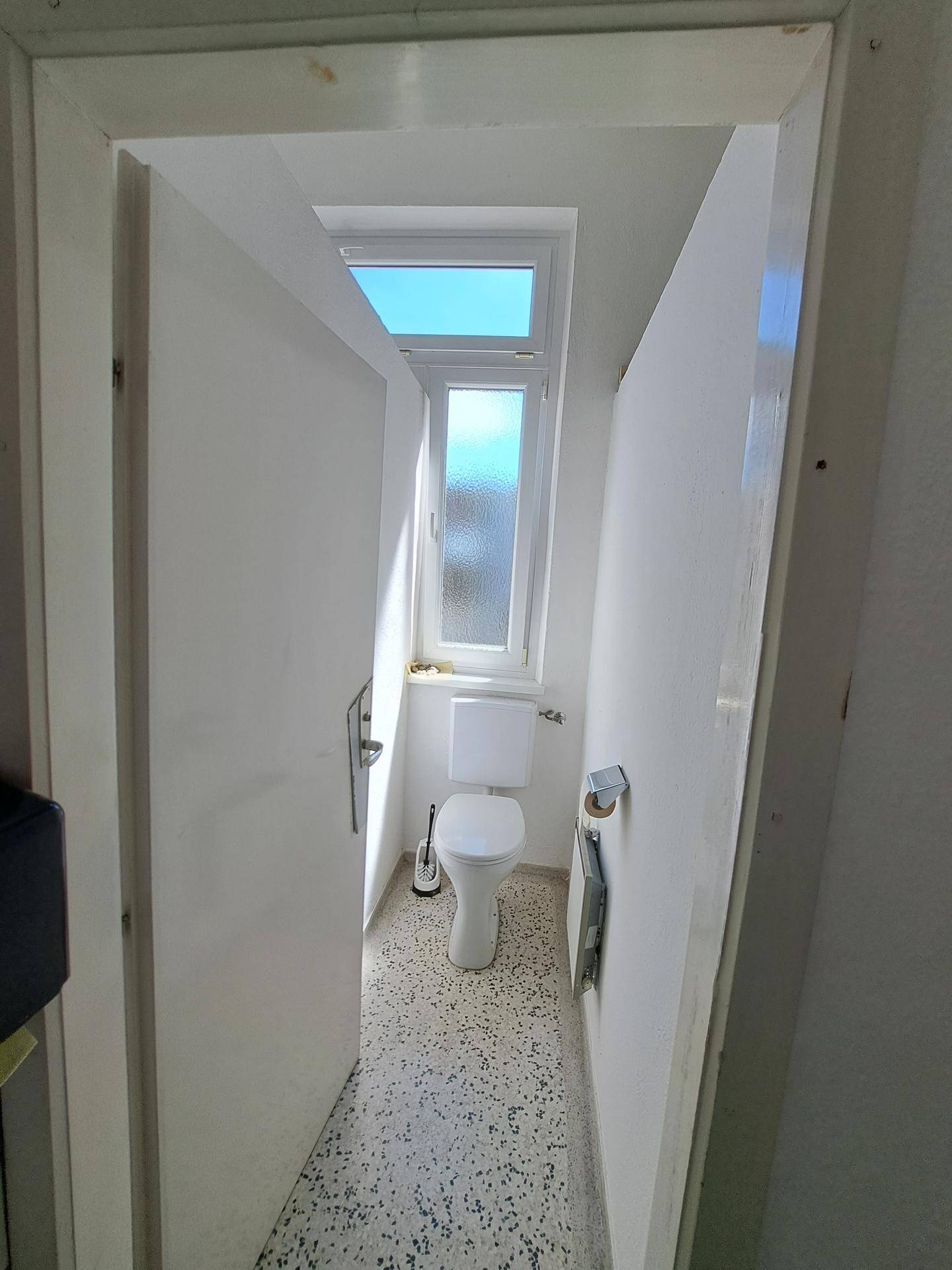Kleiner Toilettenraum mit weißem WC, Terrazzoboden und einem Fenster für Tageslicht.