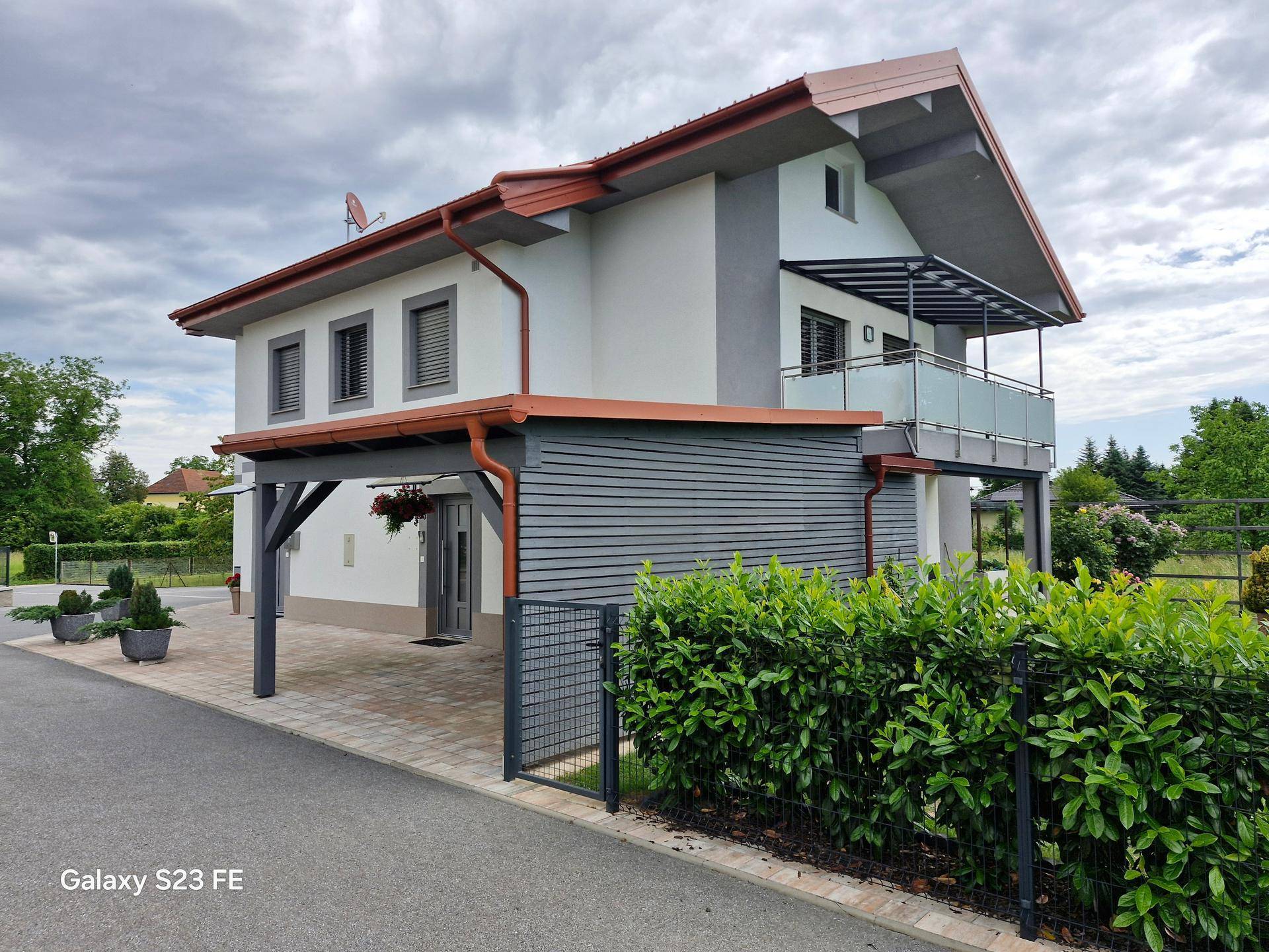 Seitenansicht des Hauses mit Carport, Balkon im Obergeschoss und gepflegter Hecke im Vordergrund.