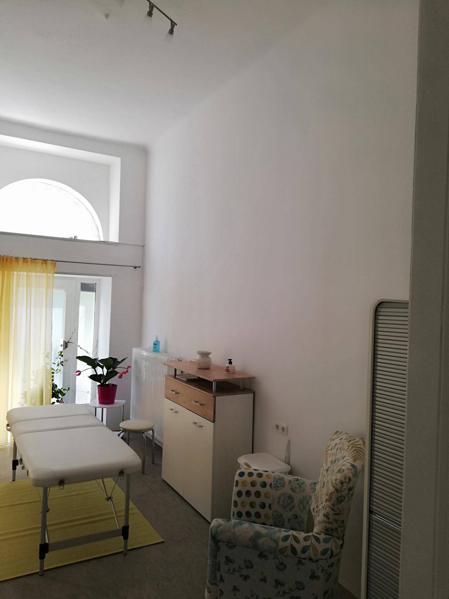 Geräumiger Behandlungsraum mit hoher Decke, einer Liege, einem Schrank und einem Fenster mit Bogen.