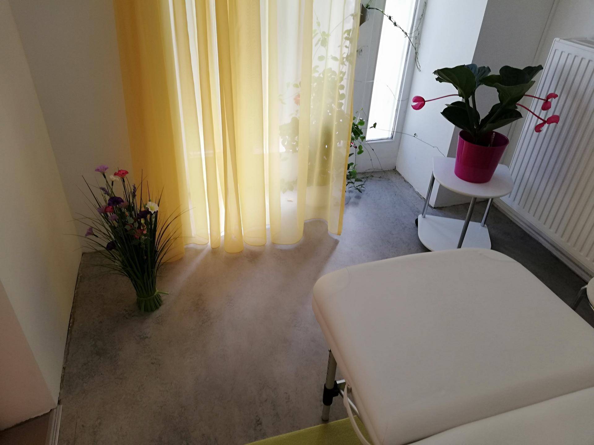Ein Behandlungsraum mit einer Liege, gelben Vorhängen vor dem Fenster und einem Heizkörper.