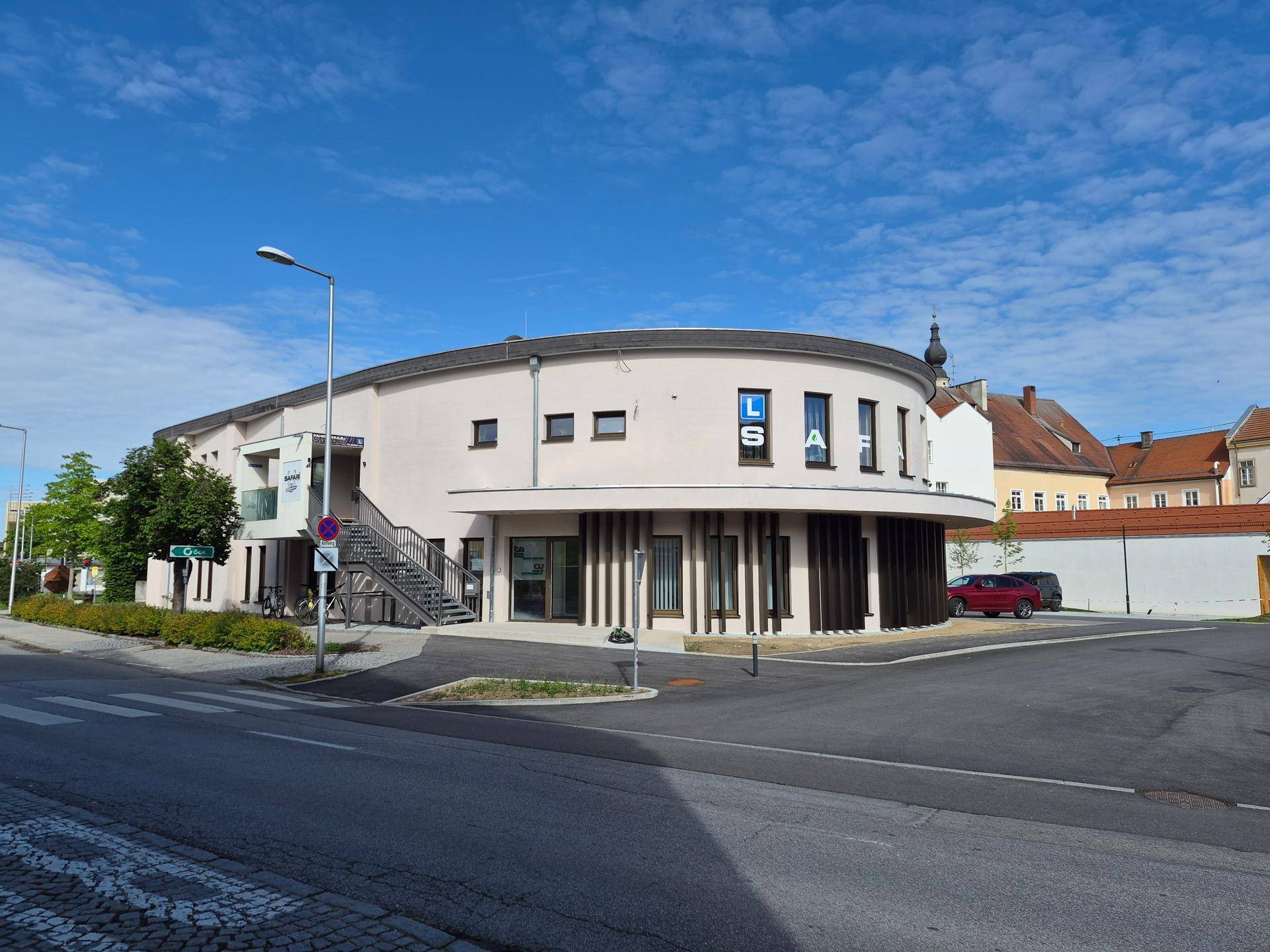 Modernes, geschwungenes Gebäude mit heller Fassade, großen Fenstern und Außentreppe an einer Straße gelegen.