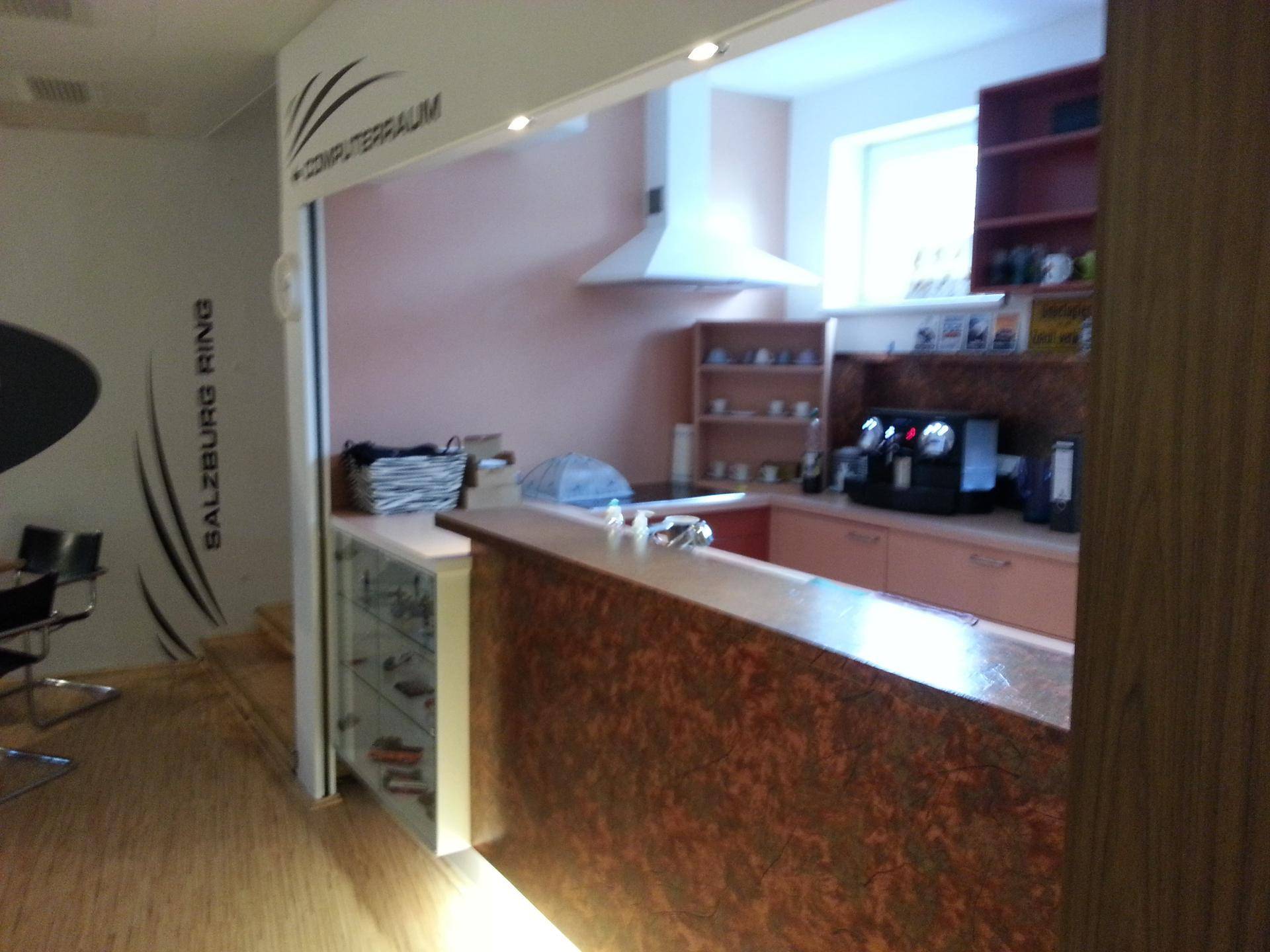 Kleine Küchenzeile mit Theke, Kaffeemaschine, Dunstabzugshaube und Fenster, integriert in einen größeren Raum.