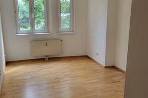 Helles Zimmer mit Holzfußboden und großen Fenstern, die eine angenehme Atmosphäre schaffen.