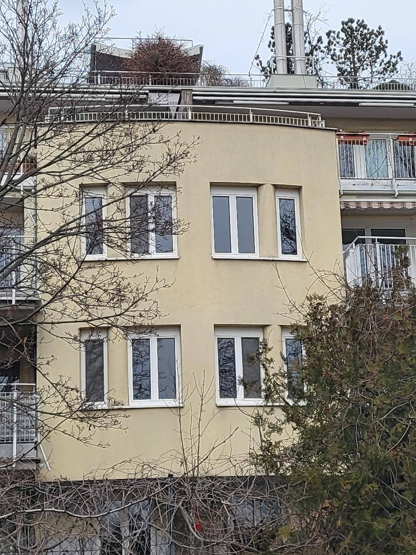 Mehrstöckiges Gebäude mit heller Fassade und mehreren Fenstern, umgeben von Bäumen.