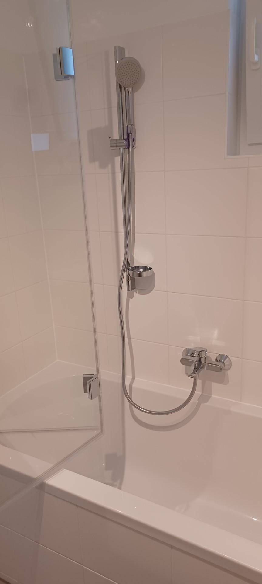 Badezimmer mit Badewanne, integrierter Dusche und Glasabtrennung, umgeben von weißen Fliesen.