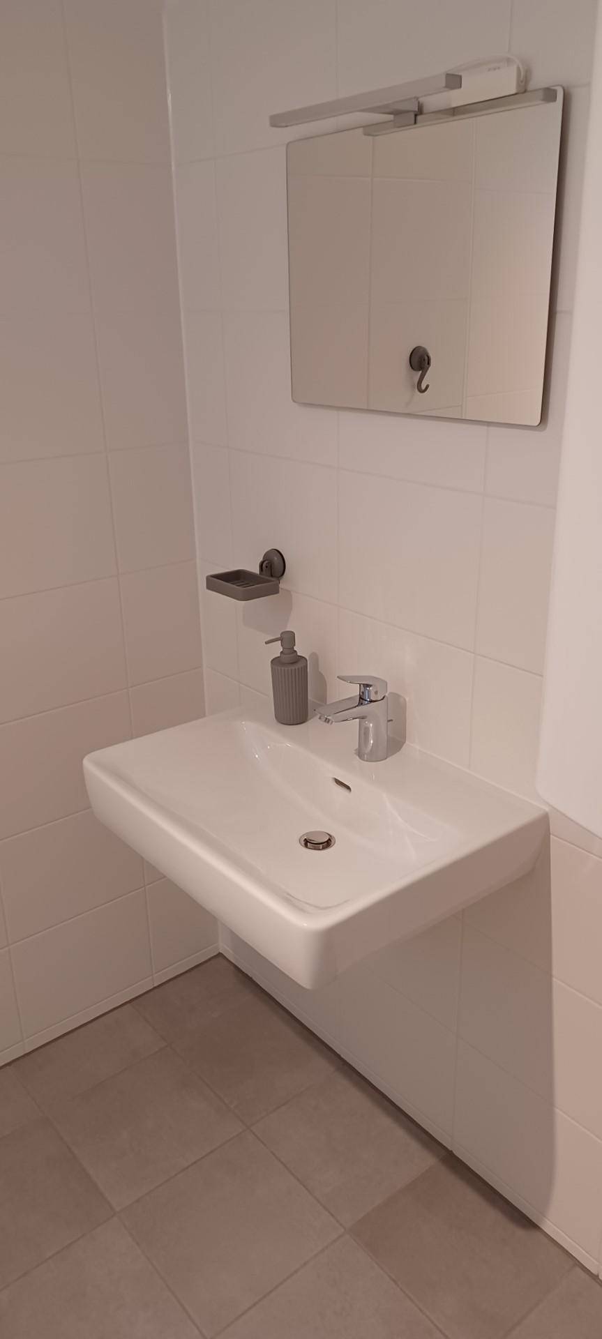 Modernes Badezimmer mit wandmontiertem Waschbecken, Spiegel und hellen Fliesen an den Wänden.