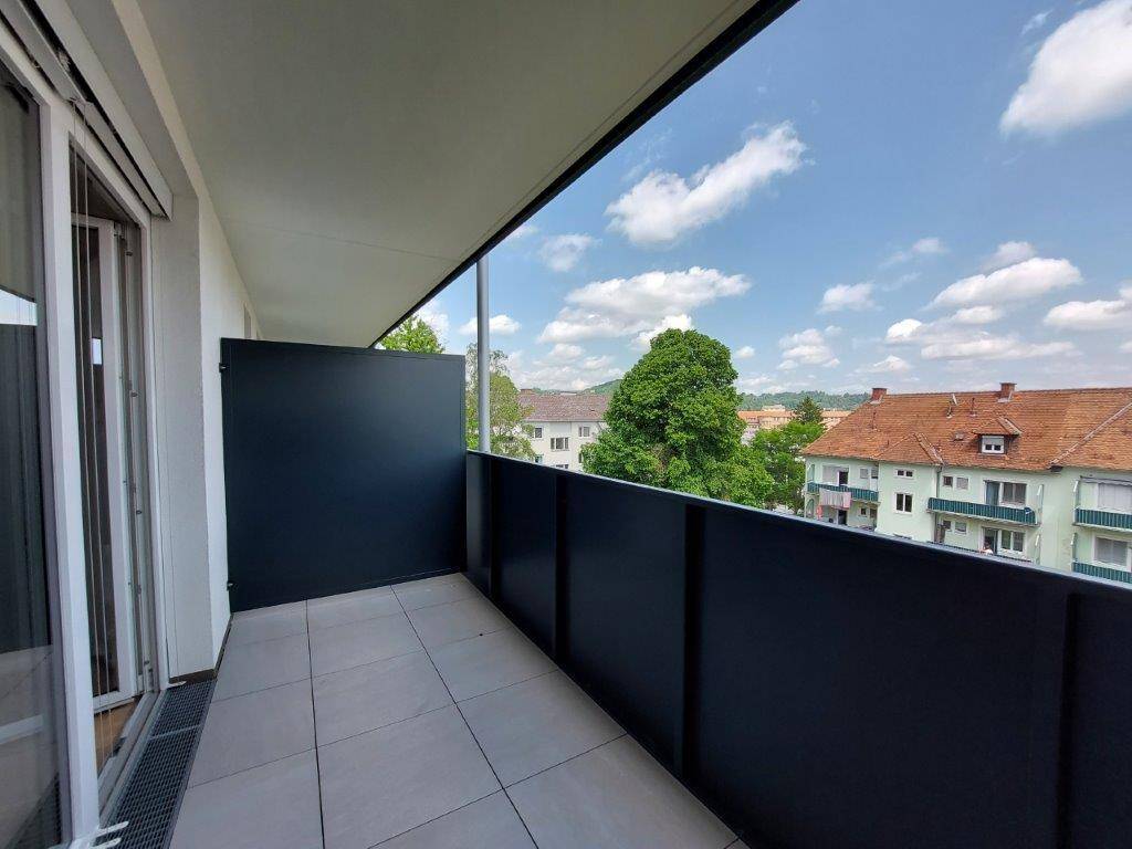 Geräumiger Balkon mit grauen Fliesen und schwarzem Geländer, bietet einen Blick auf die Umgebung und Bäume.