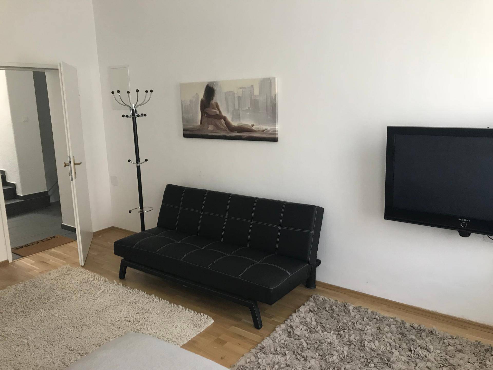 Wohnbereich mit schwarzem Schlafsofa, Wandfernseher und Garderobenständer, hell und funktional.
