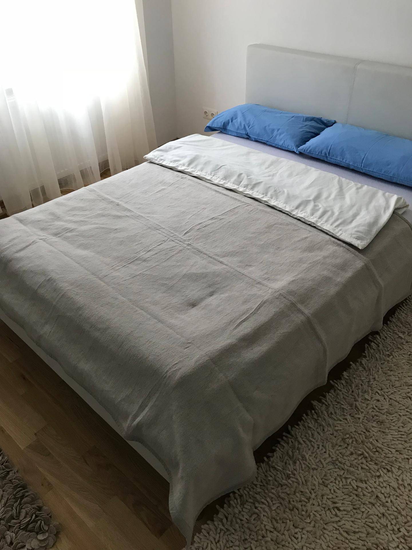 Helles Schlafzimmer mit Doppelbett, Teppich und viel Tageslicht durch das große Fenster.