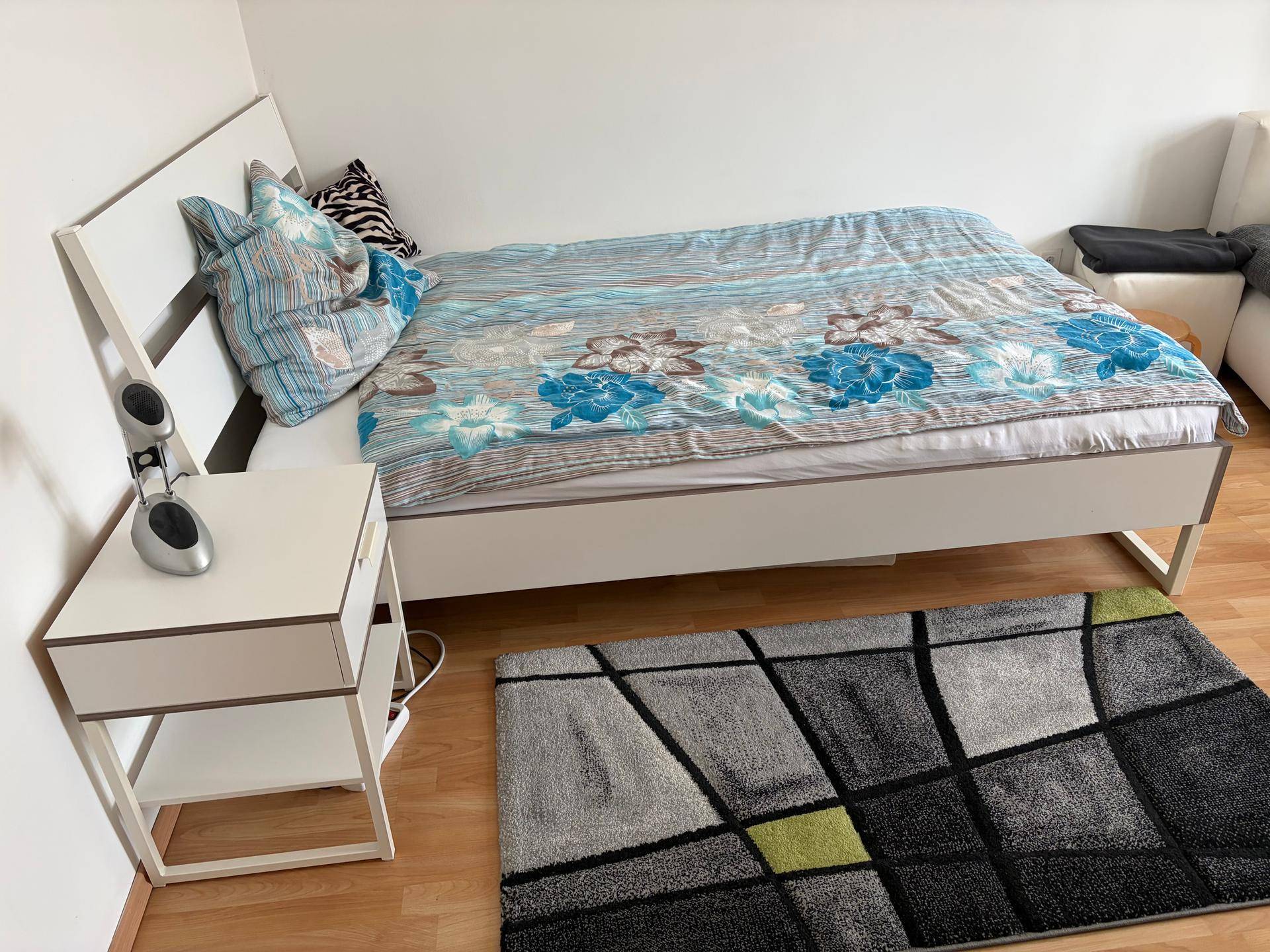 Schlafzimmer mit weißem Bett, Nachttisch und modernem Teppich auf Laminatboden.