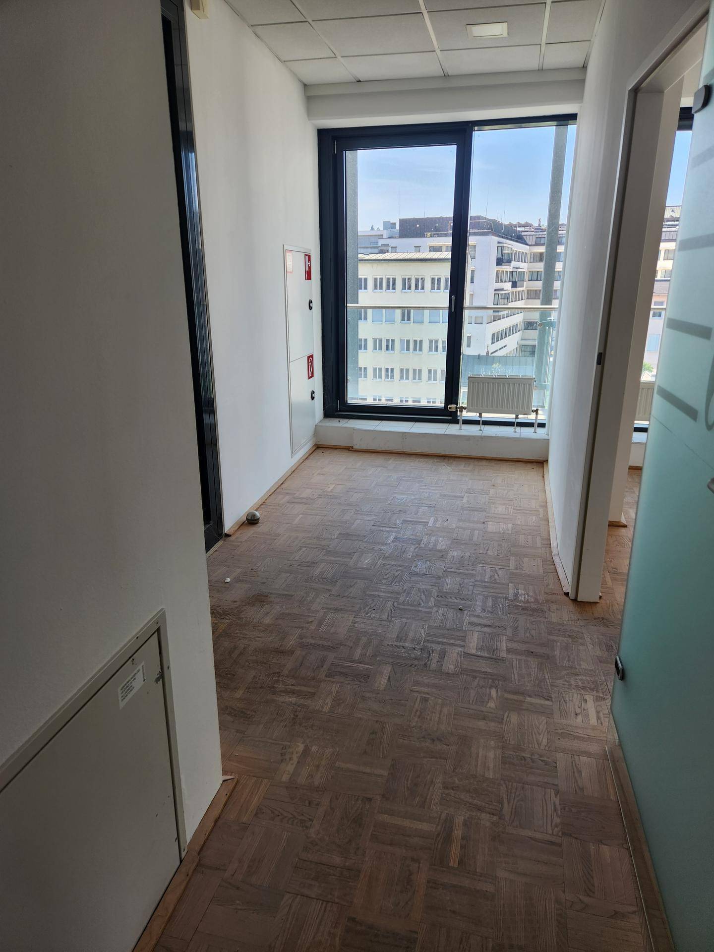 Leeres Büro mit Fischgrätparkett und großen Fenstern, die viel Tageslicht hereinlassen.