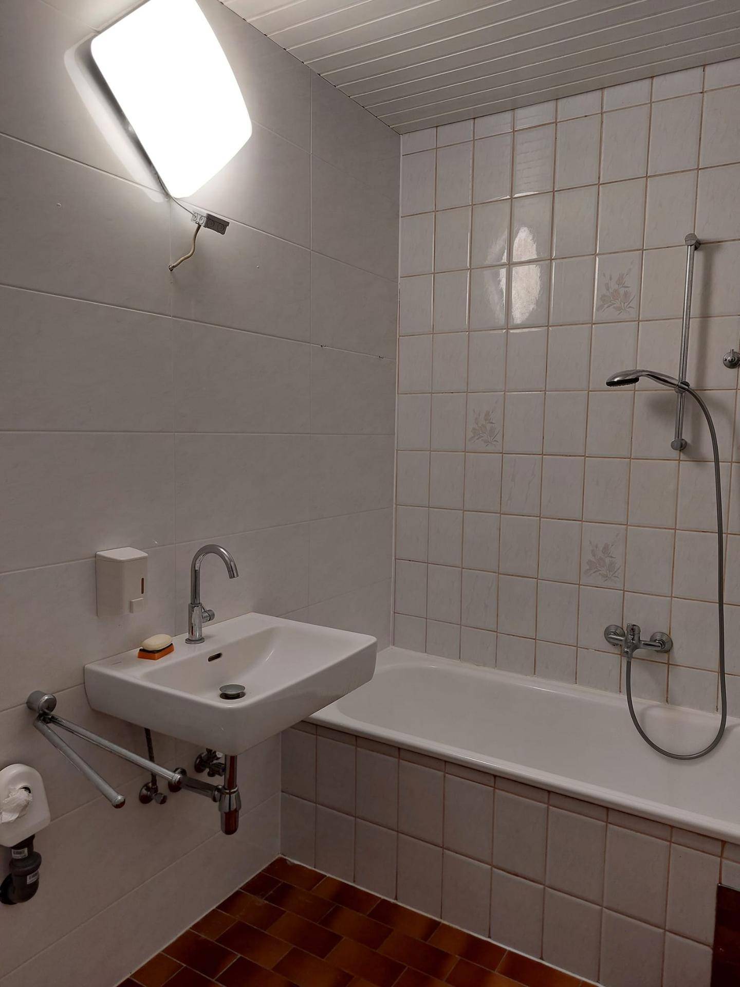 Badezimmer mit weißen Fliesen, Badewanne mit Duschfunktion, Waschbecken und braunem Fliesenboden.