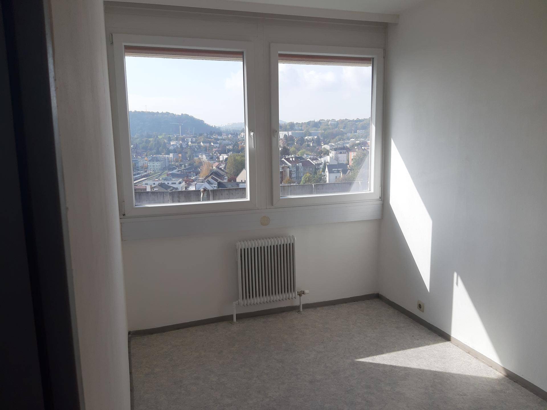 Leerer Raum mit hellem Boden, weißen Wänden und zwei Fenstern mit weitem Blick über die Umgebung.