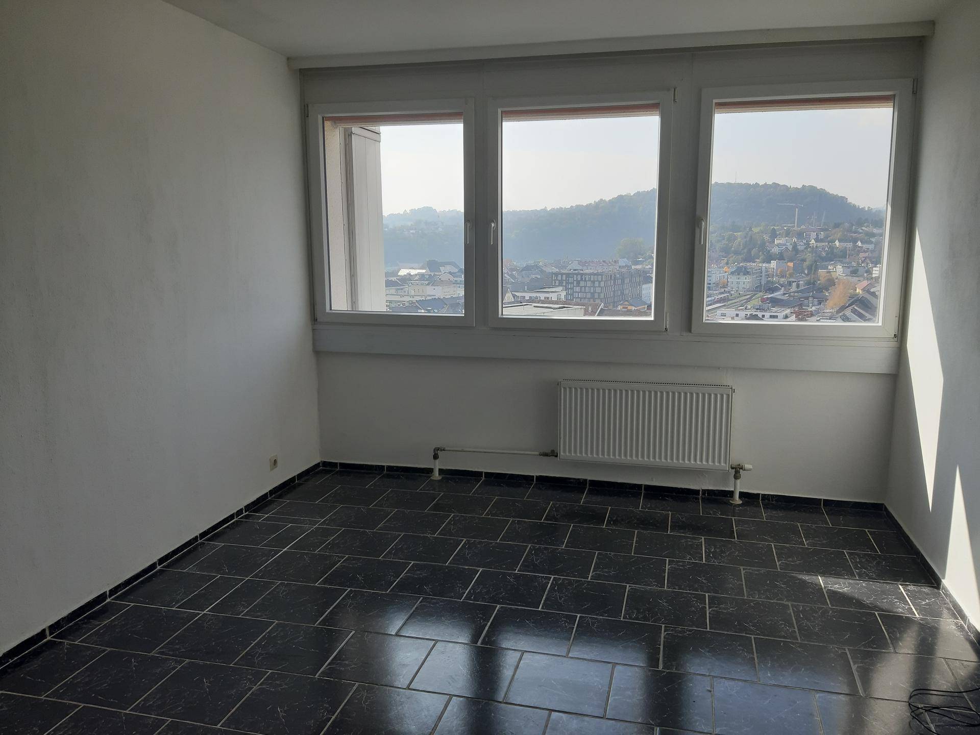Leerer Raum mit dunklen Fliesen, weißen Wänden und drei Fenstern mit Blick auf die Stadt und Landschaft.