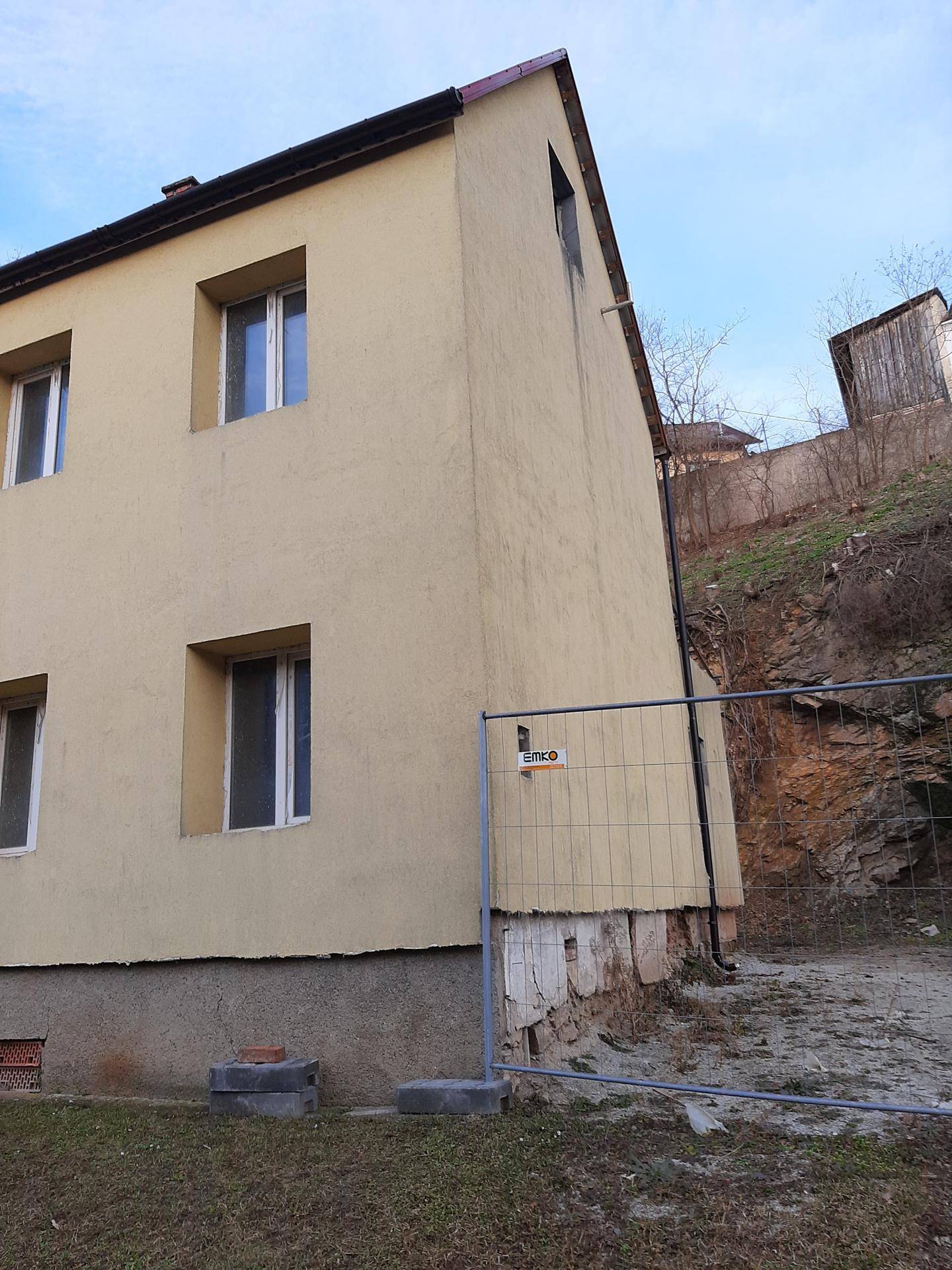 Ecke des gelben Gebäudes mit Bauzaun und felsigem Hang im Hintergrund.