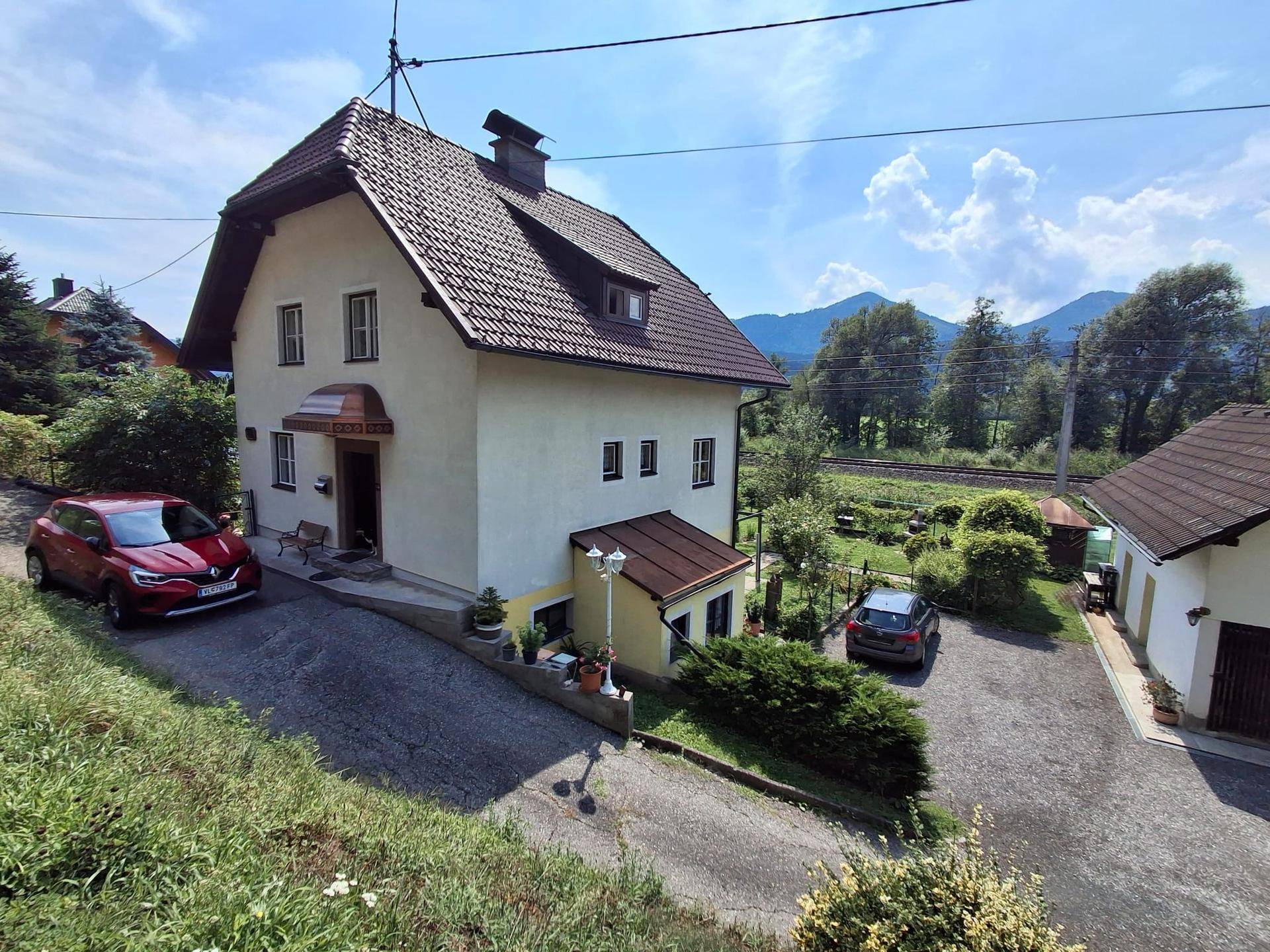 Haus mit zwei Autos in der Einfahrt, umgeben von grüner Landschaft und Bergen.