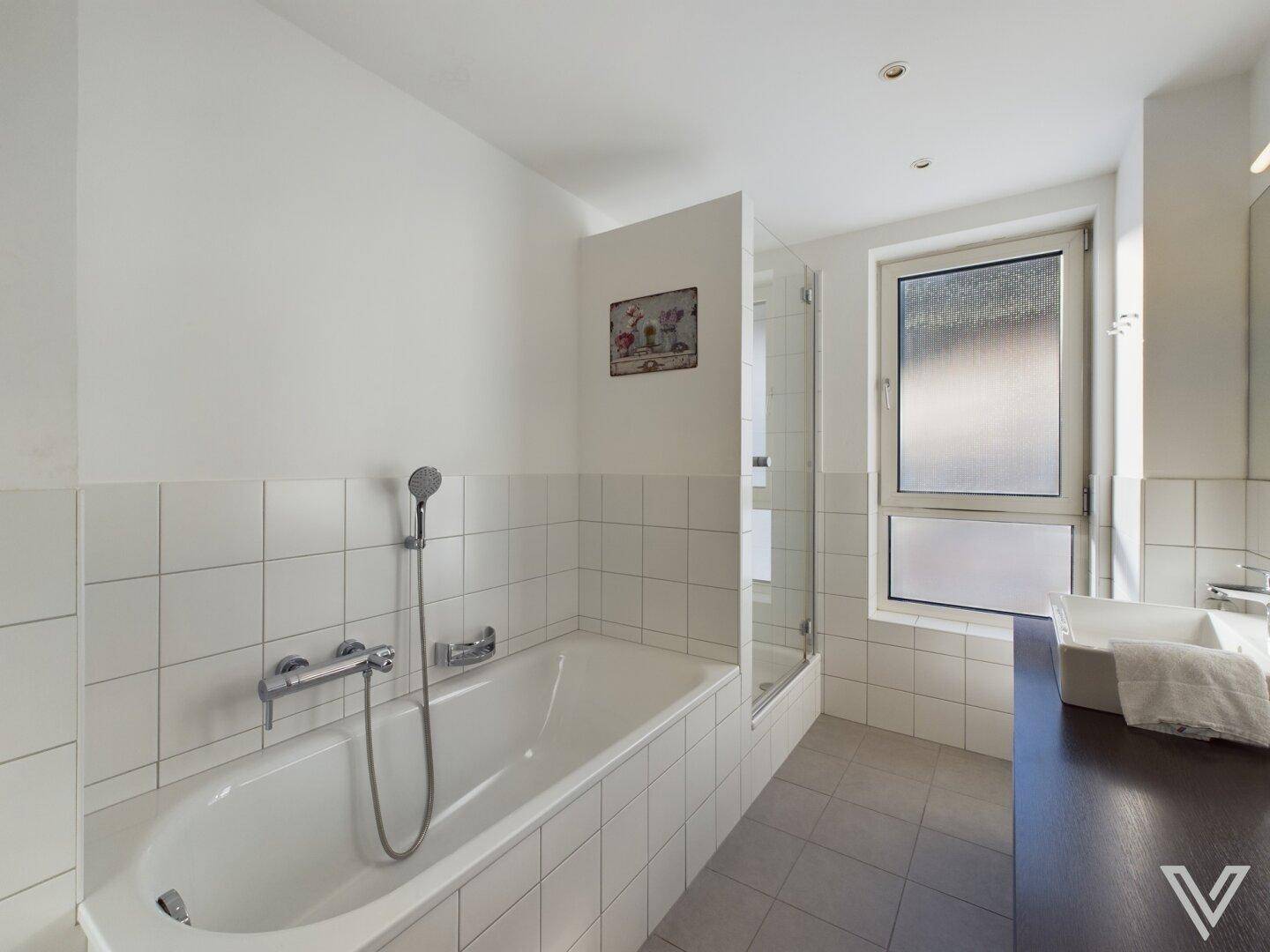 Modernes Badezimmer mit Badewanne, Dusche und Fenster, hell gefliest in Weiß.