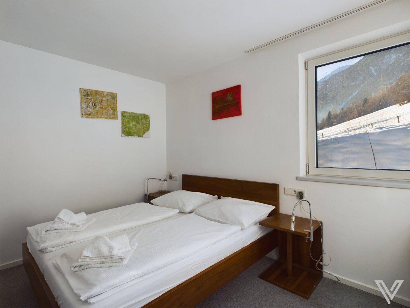 Gemütliches Schlafzimmer mit Holzbett und Fenster mit Blick auf die Berge und verschneite Landschaft.