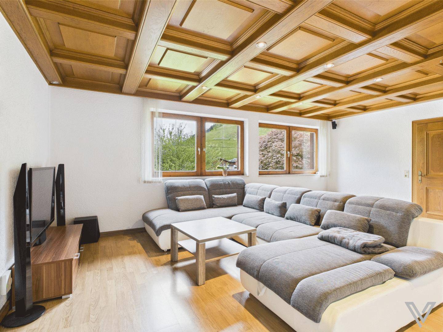 Geräumiges Wohnzimmer mit großer Eckcouch, Holzkassettendecke und Blick ins Grüne.