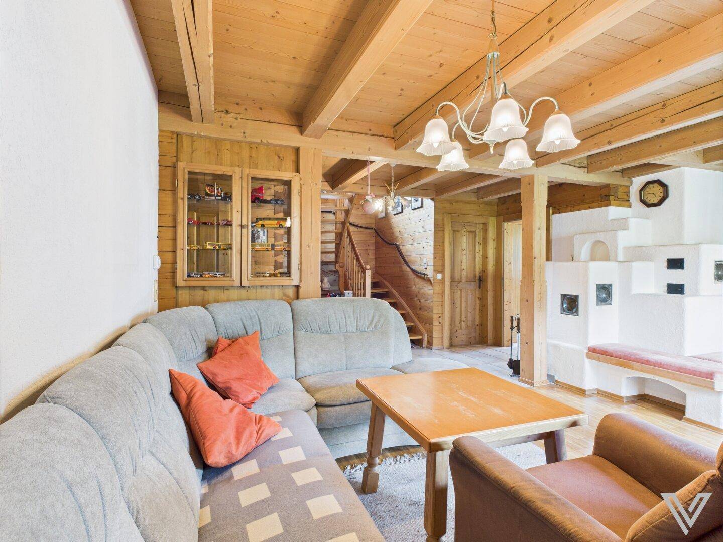 Wohnzimmer im Landhausstil mit bequemer Couch, Holzbalkendecke und traditionellem Kachelofen.
