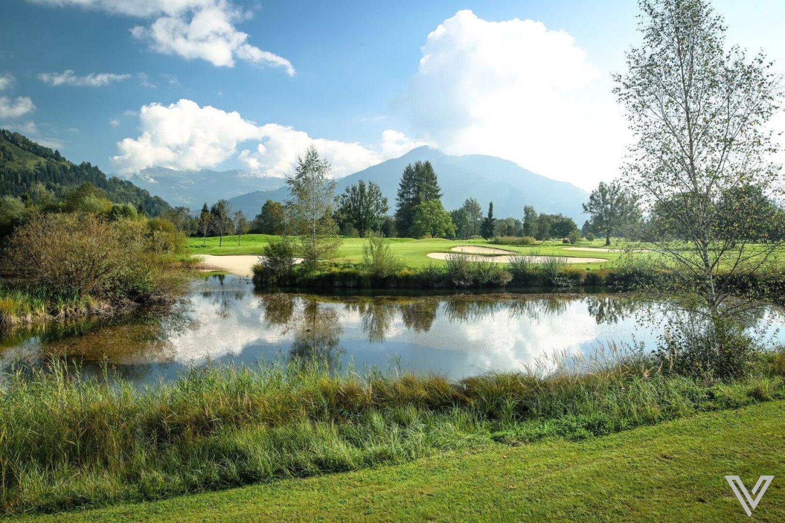Idyllischer Teich mit Spiegelungen des Himmels und der Berge, umgeben von Grünflächen.