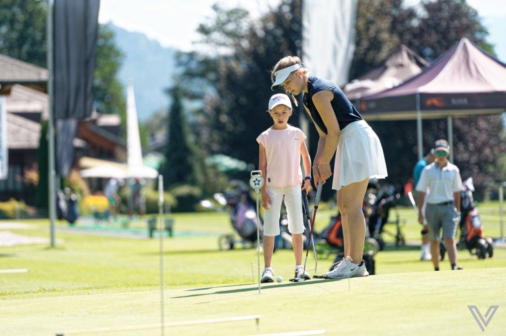 Golftraining für Kinder auf dem Putting Green mit einer Trainerin und Bergkulisse.
