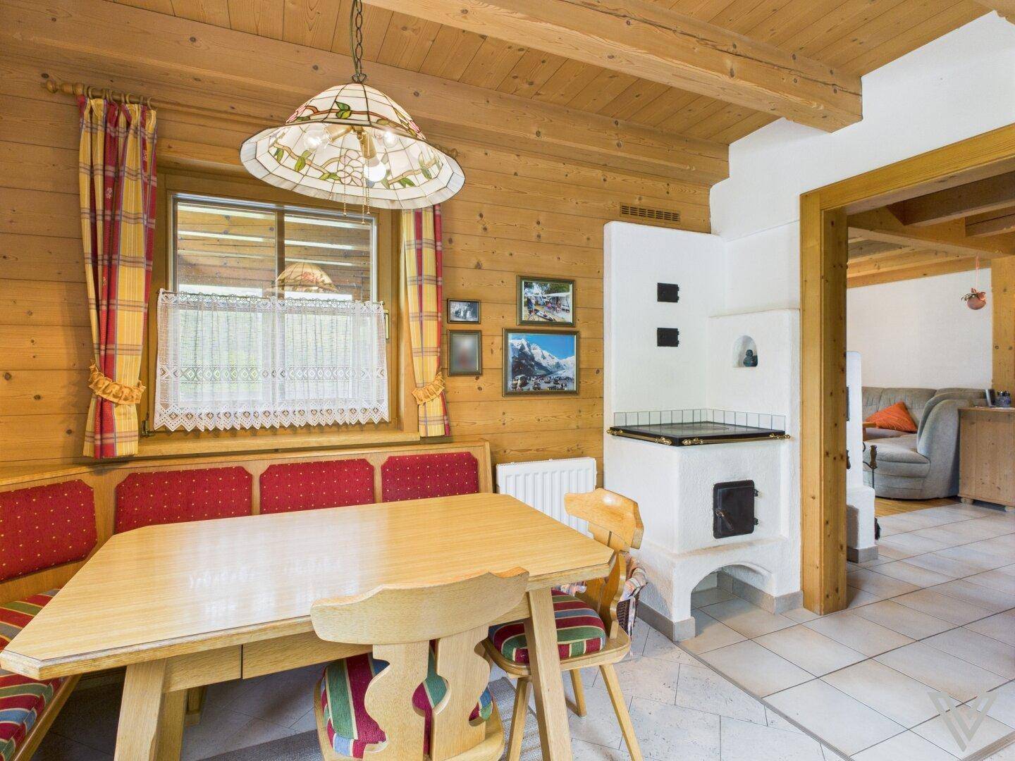 Gemütlicher Essbereich mit Eckbank, Holztisch und traditionellem Kachelofen im Hintergrund.