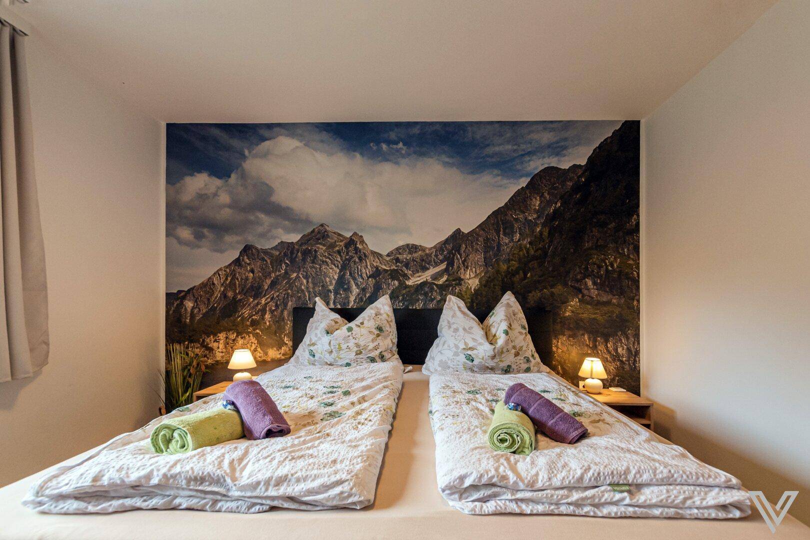 Schlafzimmer mit Doppelbett, beeindruckendem Bergblick-Wandbild und Holzboden.