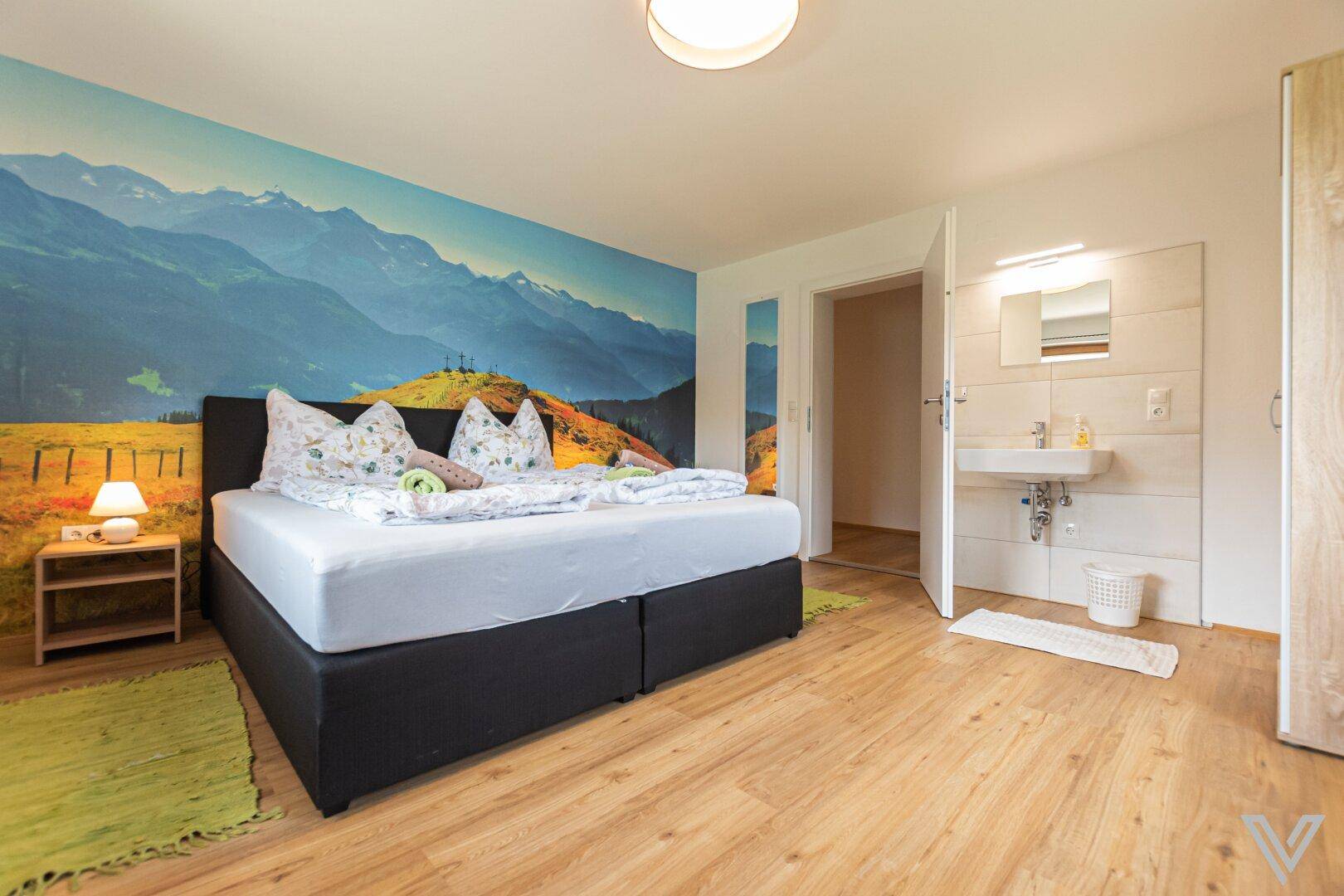 Schlafzimmer mit Bergblick-Wandbild und angrenzendem Badezimmer mit Waschbecken.