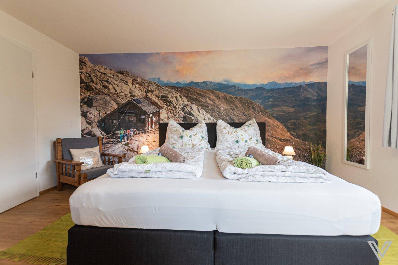 Schlafzimmer mit Doppelbett, Bergblick-Wandbild und rustikalem Sessel zum Entspannen.